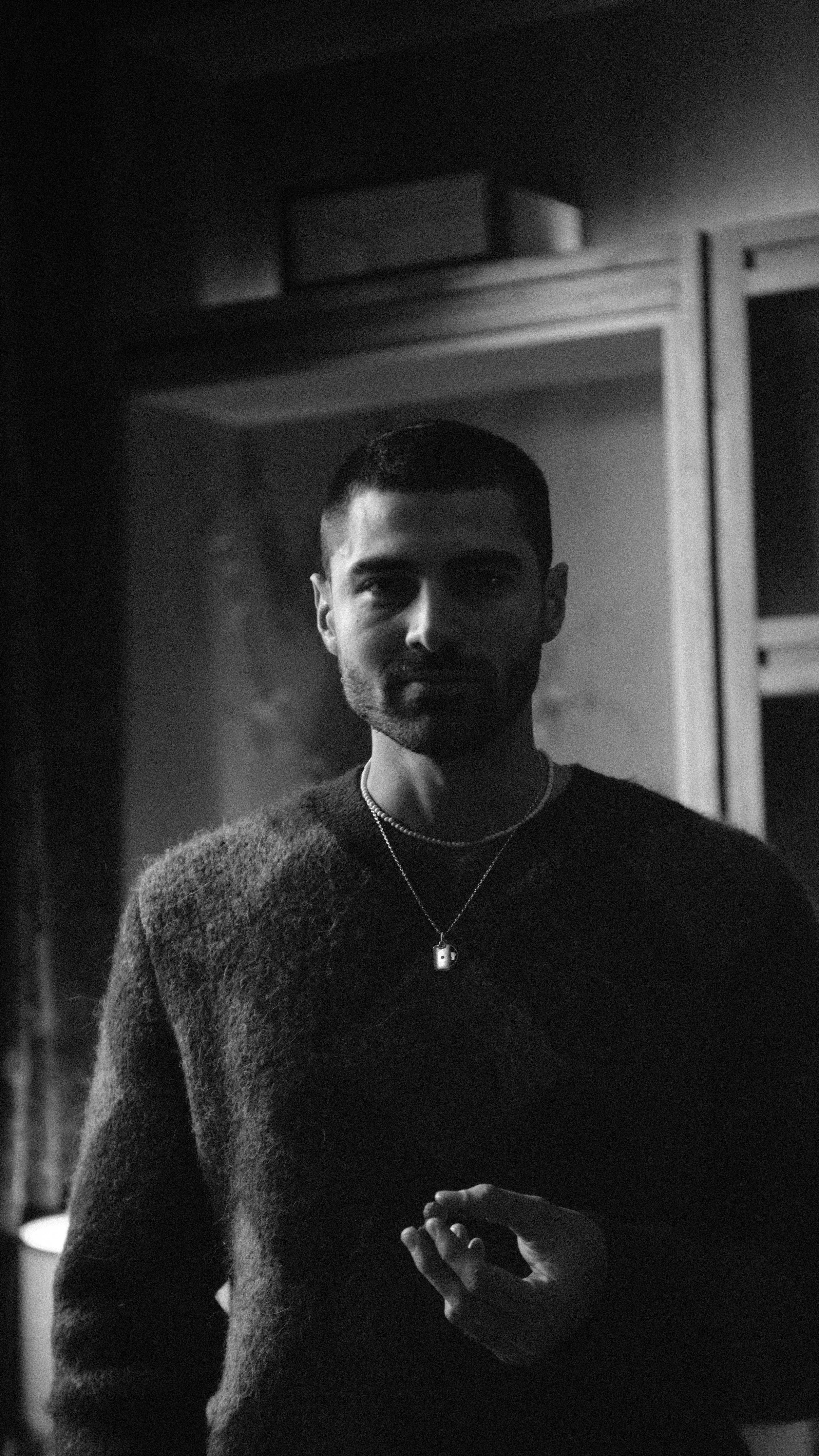 A black and white photo of a young man with short dark hair and a beard, wearing a dark sweater and layered necklaces, standing indoors in front of a cabinet, looking at the camera with a subtle smile.