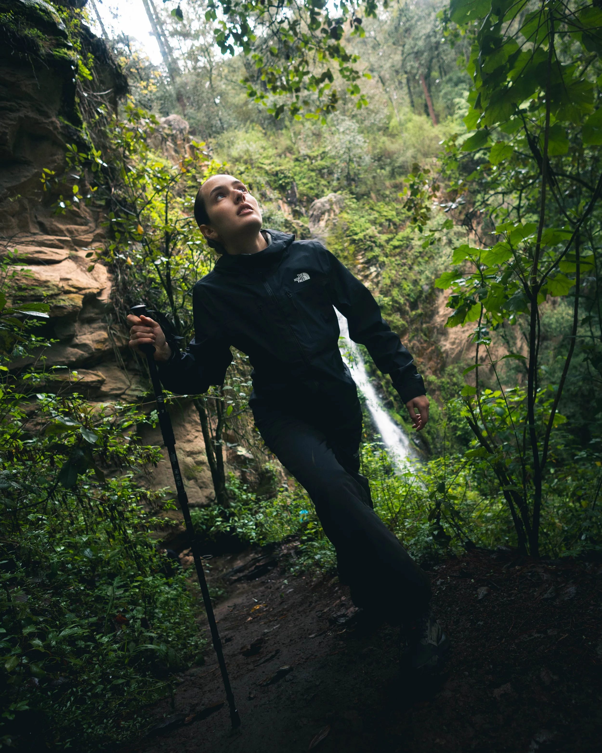 A woman in black outdoor gear holding a trekking pole, looking up in a lush green forest with a waterfall in the background.