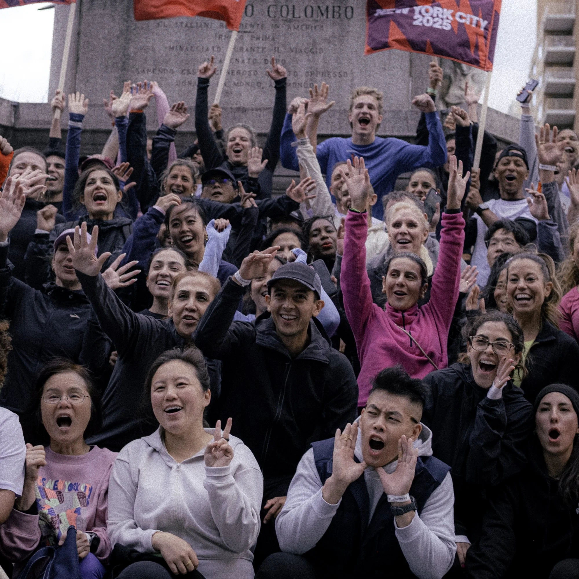 A large group of diverse people are cheering, smiling, and showing excitement at a public gathering, with some holding flags, in front of a monument.