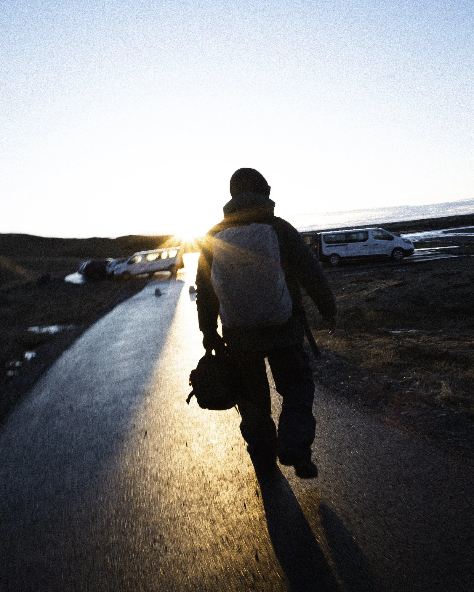 Person walking on a wet road during sunset with parked cars on side.