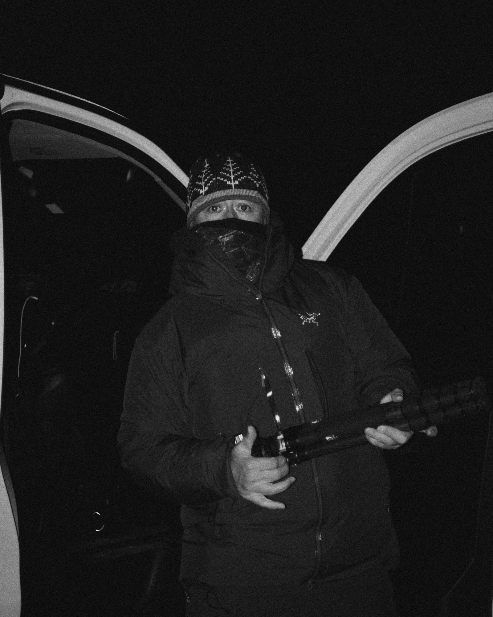 Person wearing winter clothing, holding a firearm, standing outside at night with vehicle doors open in the background.