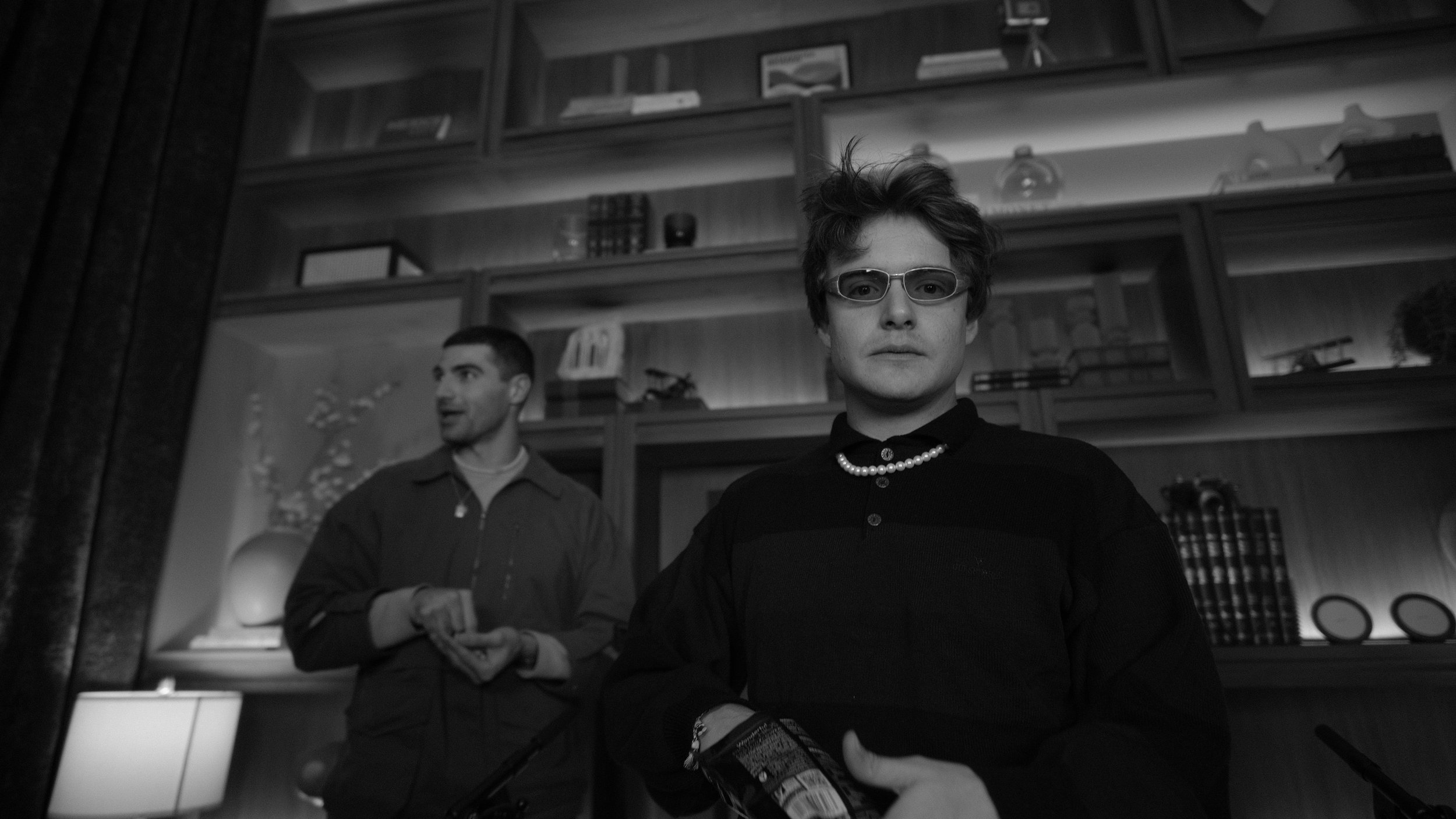 A man wearing sunglasses and a pearl necklace stands in the foreground, while another man holding a drink stands in the background in a room with wooden shelves and books.