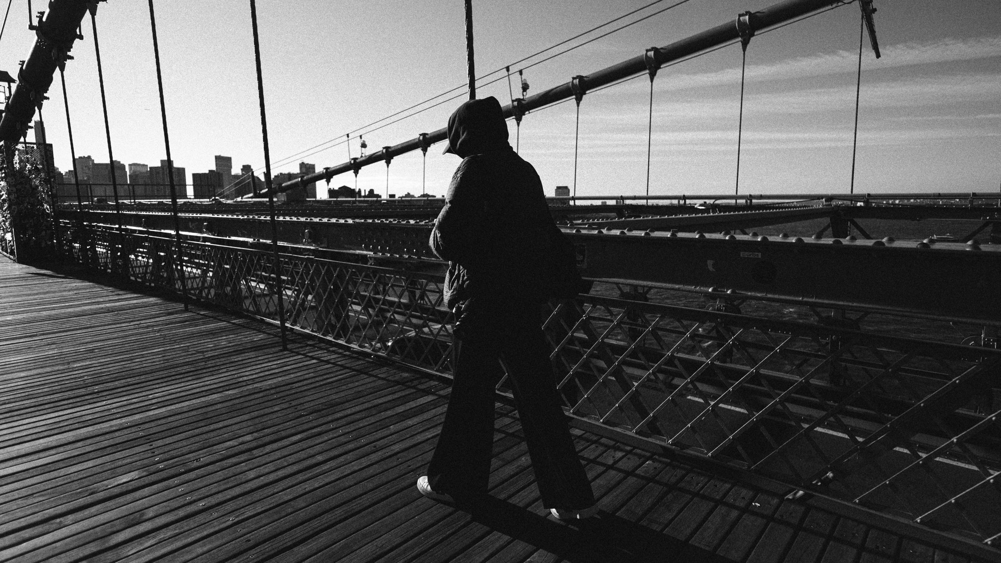 A person in dark clothing with a hood standing on a bridge, looking out over the city skyline in the distance.