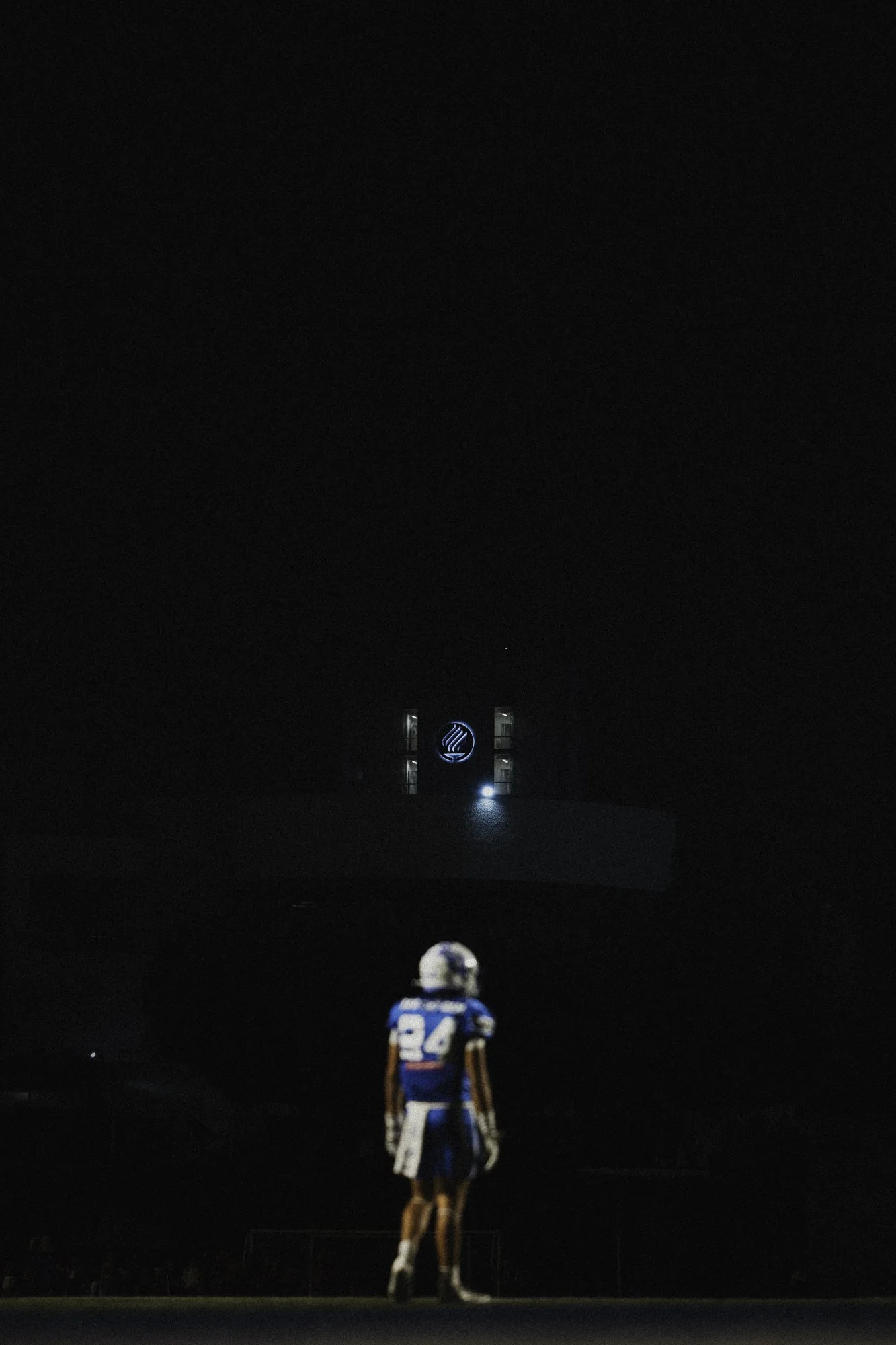 A football player wearing a jersey with the number 24 stands on a field at night, facing a dark sky with a lit building and logo in the background.