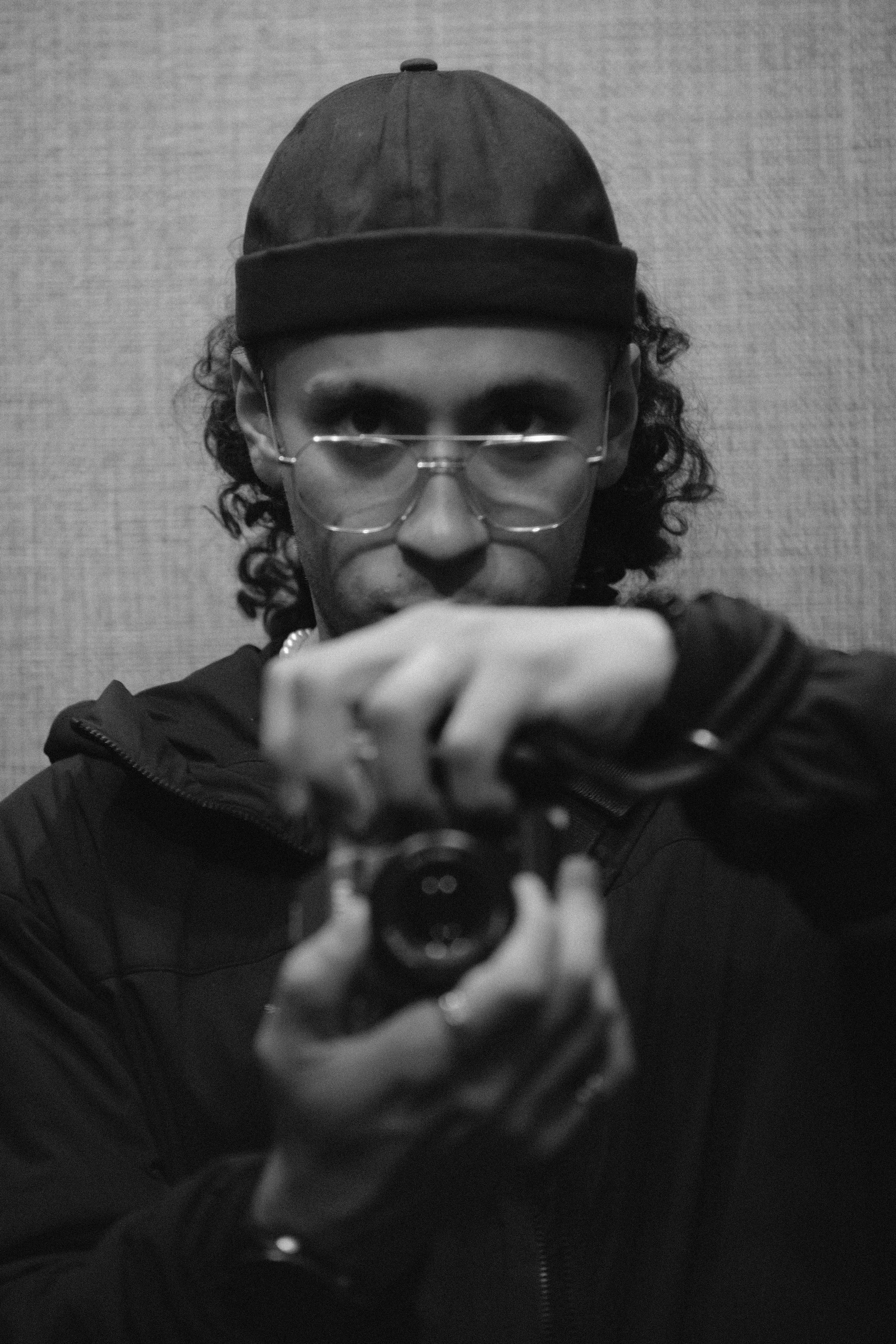 A person with curly hair, wearing glasses, a hat, and a jacket, taking a selfie with a camera in a mirror.