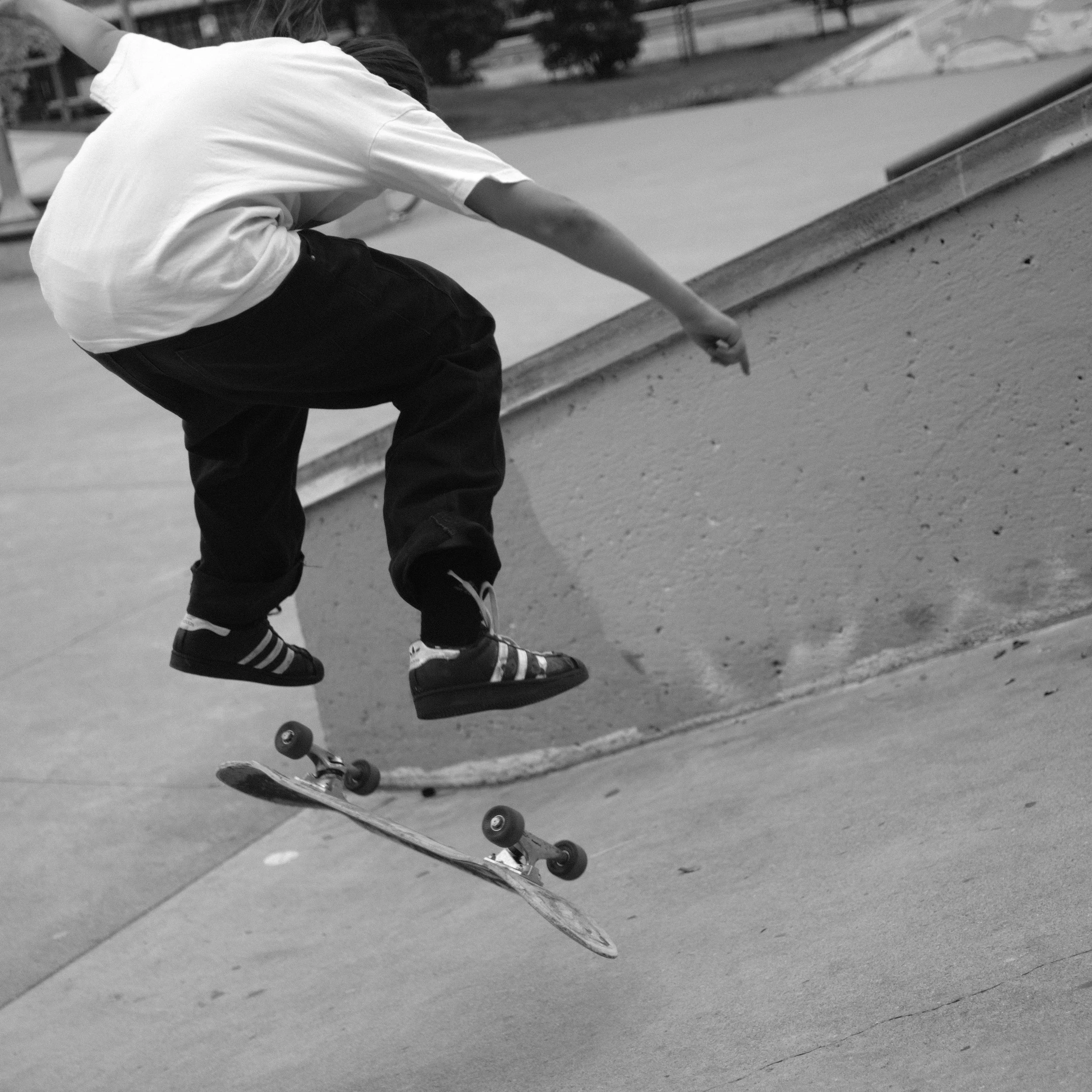 A young skateboarder in a white t-shirt, black baggy pants, and sneakers, performing a trick at a skate park on a skateboard, in black and white.