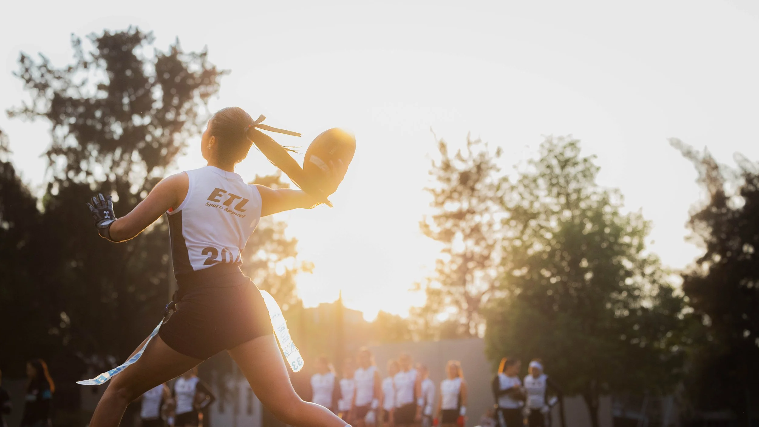 A female football player with a ponytail, wearing a white jersey with 'ETL' on the back, is throwing a football during a game at sunset, with other players in the background.