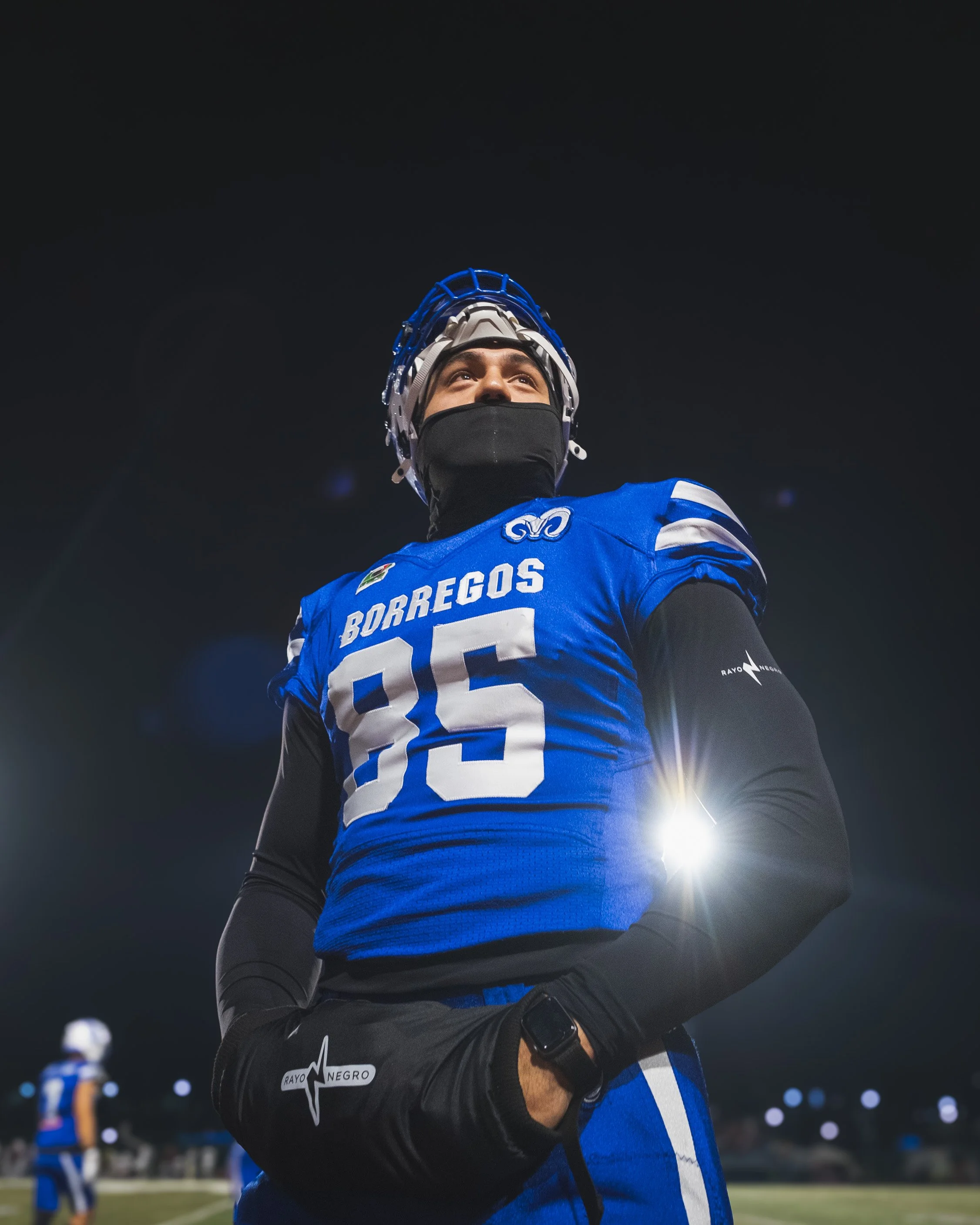 A football player in a blue jersey with the number 85 and the name Borregos, wearing a helmet and black face mask, standing on a field at night, with stadium lights and other players in the background.
