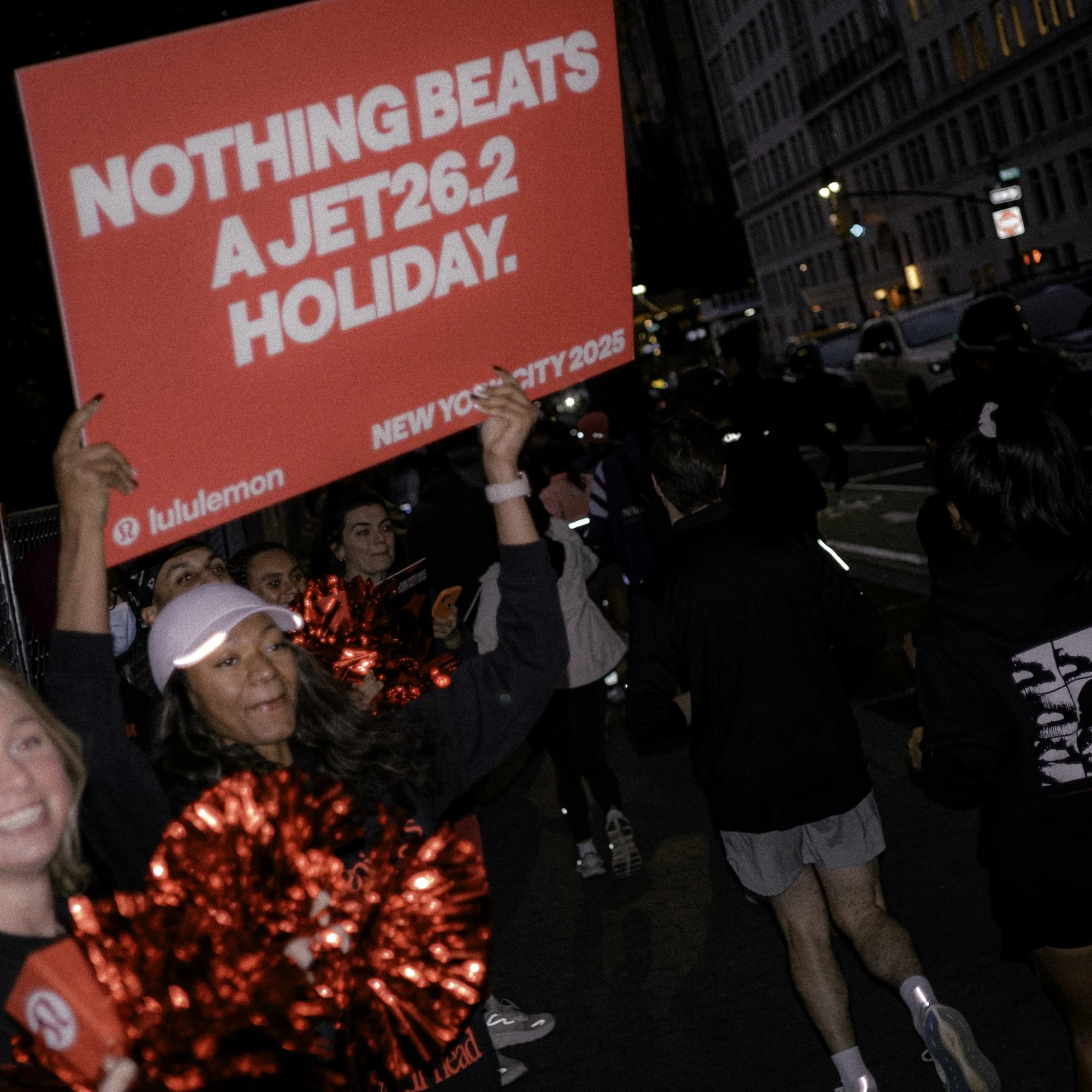 A group of people participating in a night marathon or running event in an urban setting, with some holding a large pink sign that reads "Nothing Beats a Jet 26.2 Holiday."