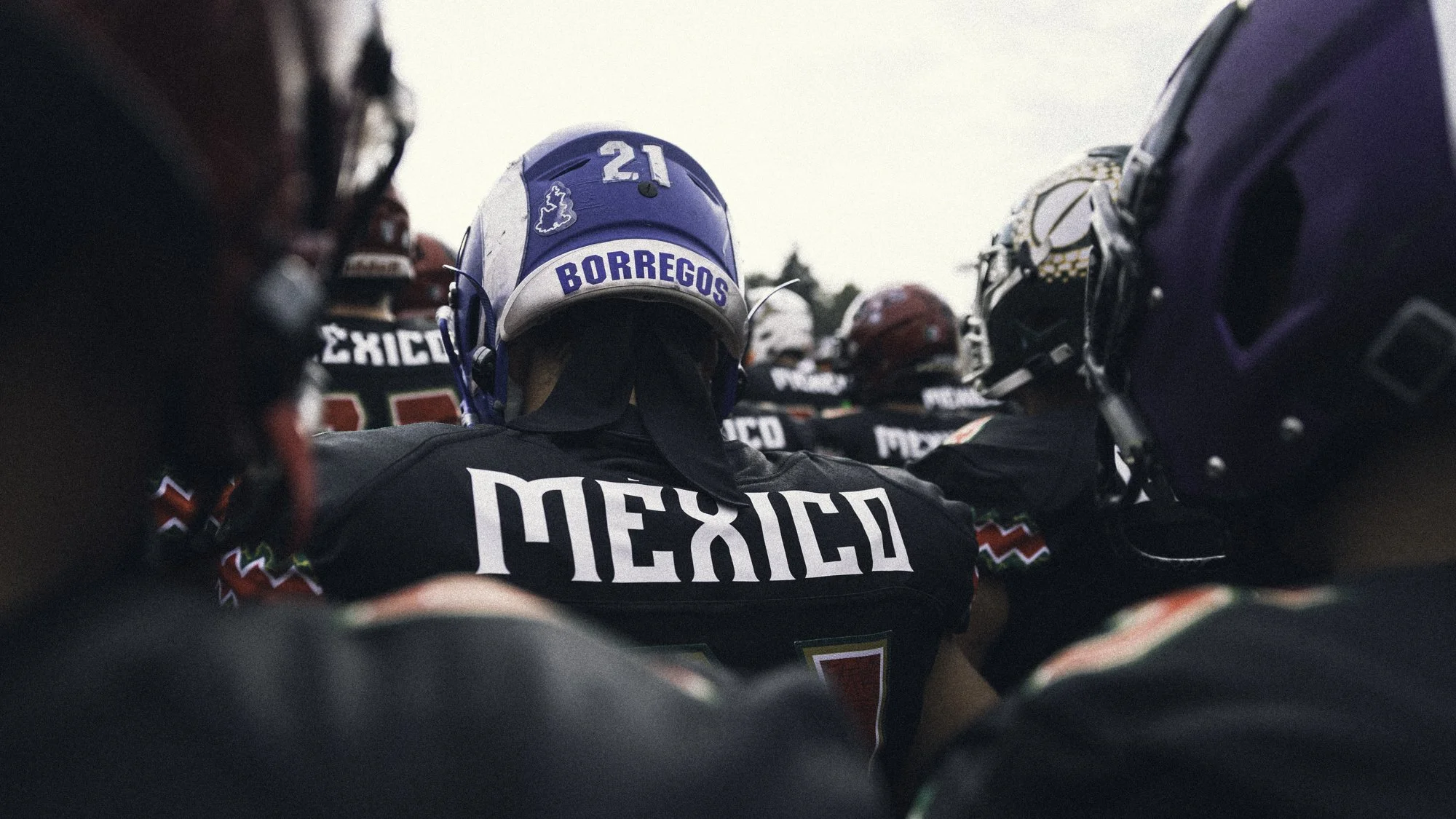 Close-up of a group of football players huddled together on the field, with one player wearing a blue helmet labeled "Borregos" and a black jersey with "México" written on the back.
