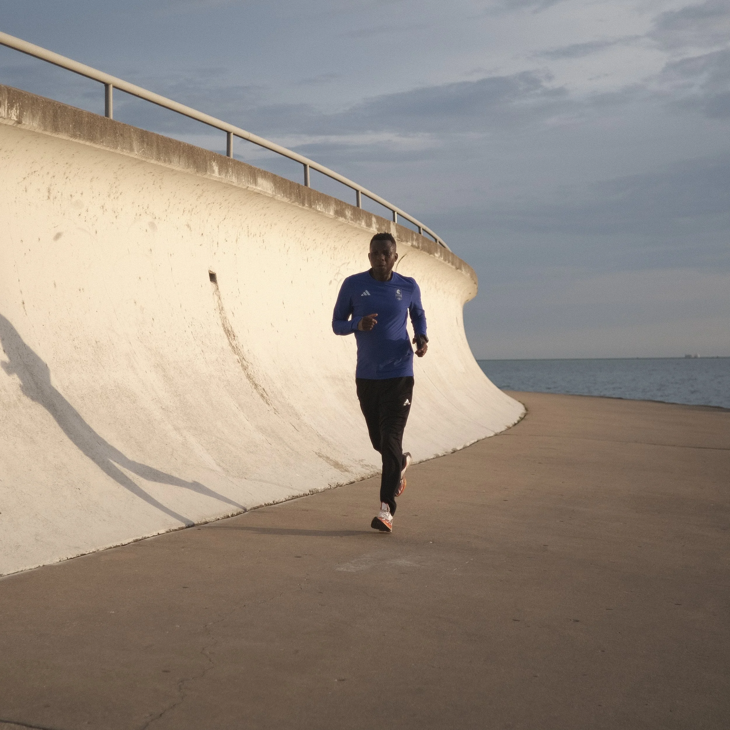 A man running along a paved path next to a curved white concrete wall near the water at dusk.