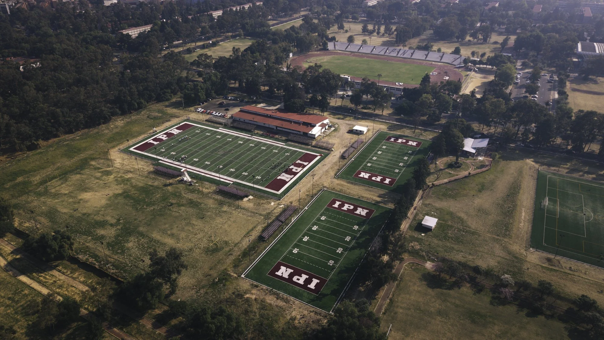 Aerial view of a sports complex with several football fields displaying the logo 'IPN' at the end zones, surrounded by trees and athletic facilities.
