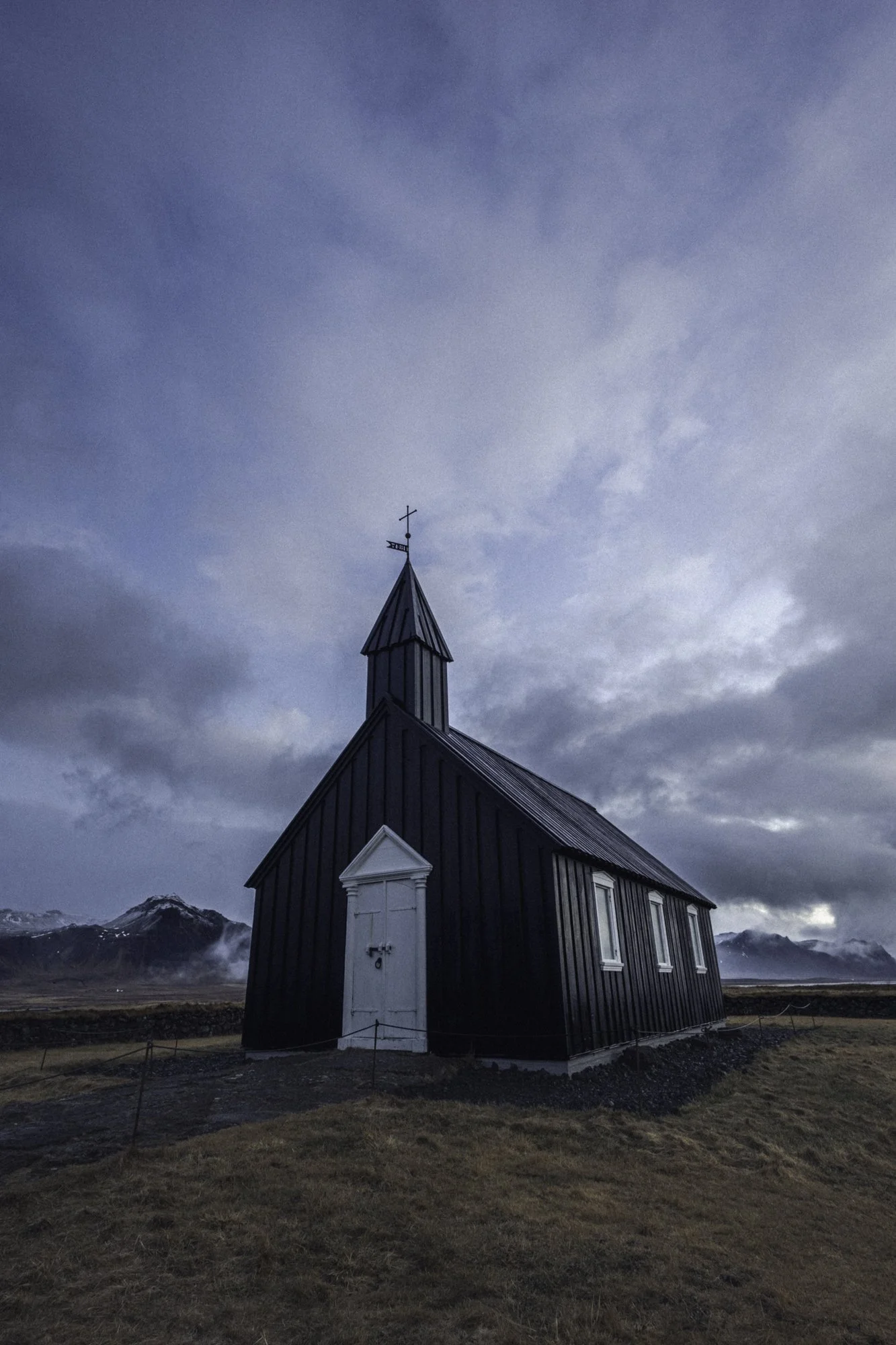 A small black church with a steeple, set against a cloudy sky and mountains in the background.