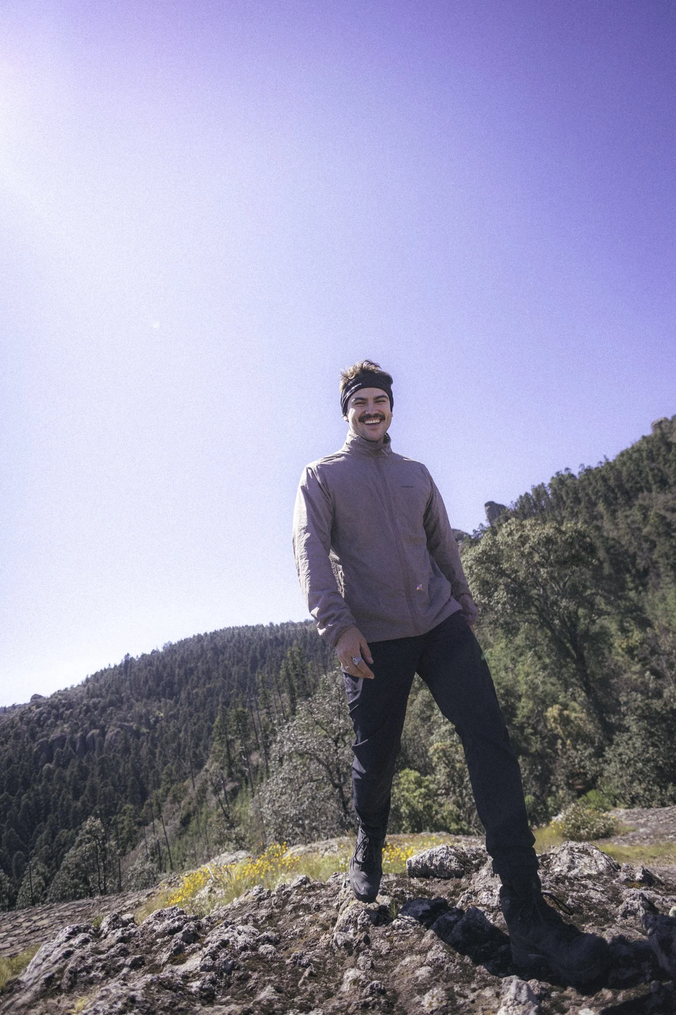 A man smiling and standing on rocks outdoors with a clear blue sky, hills, and trees in the background.