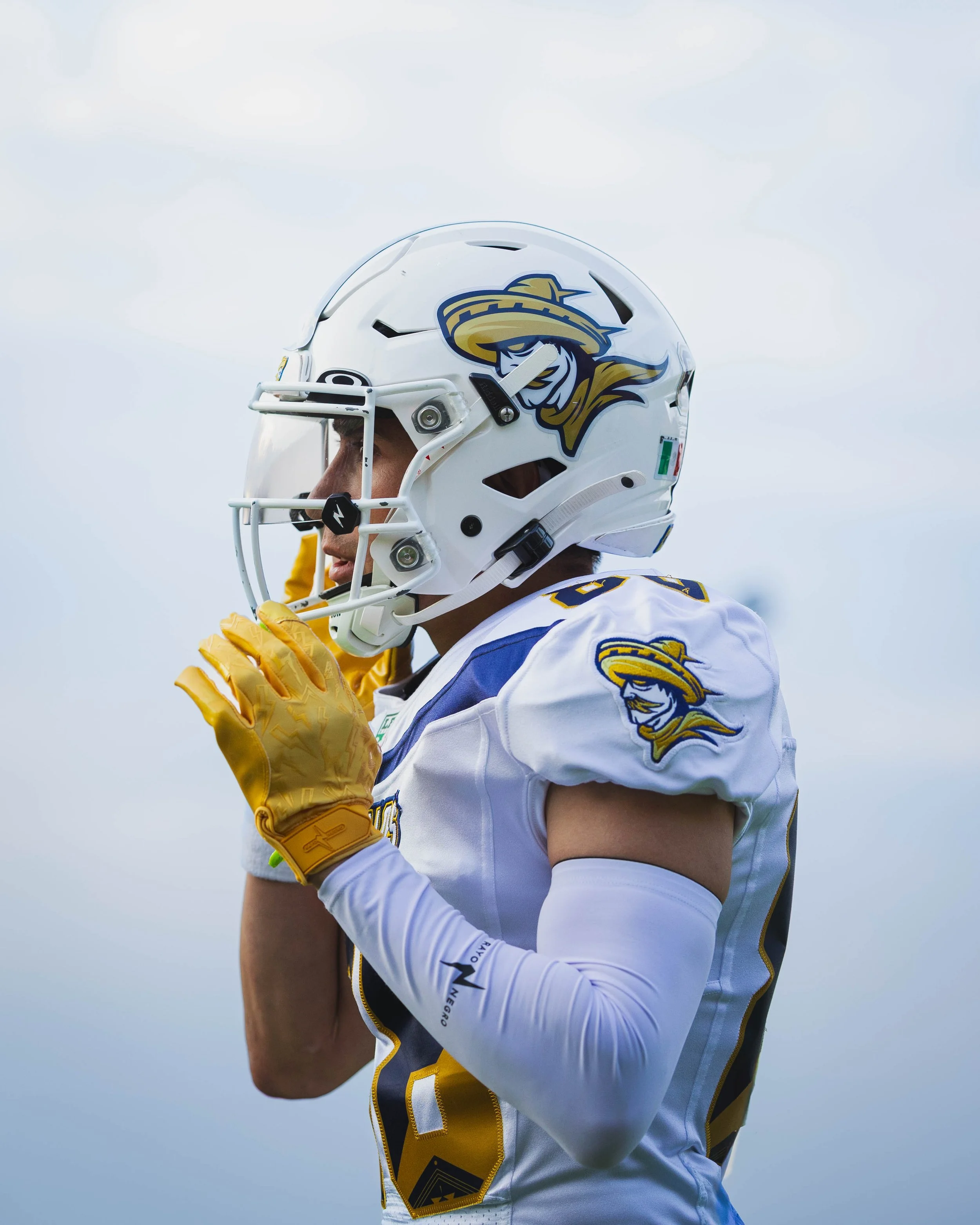 American football player in a white and blue jersey with yellow accents, wearing a helmet with a shark logo and yellow gloves, standing on the field.