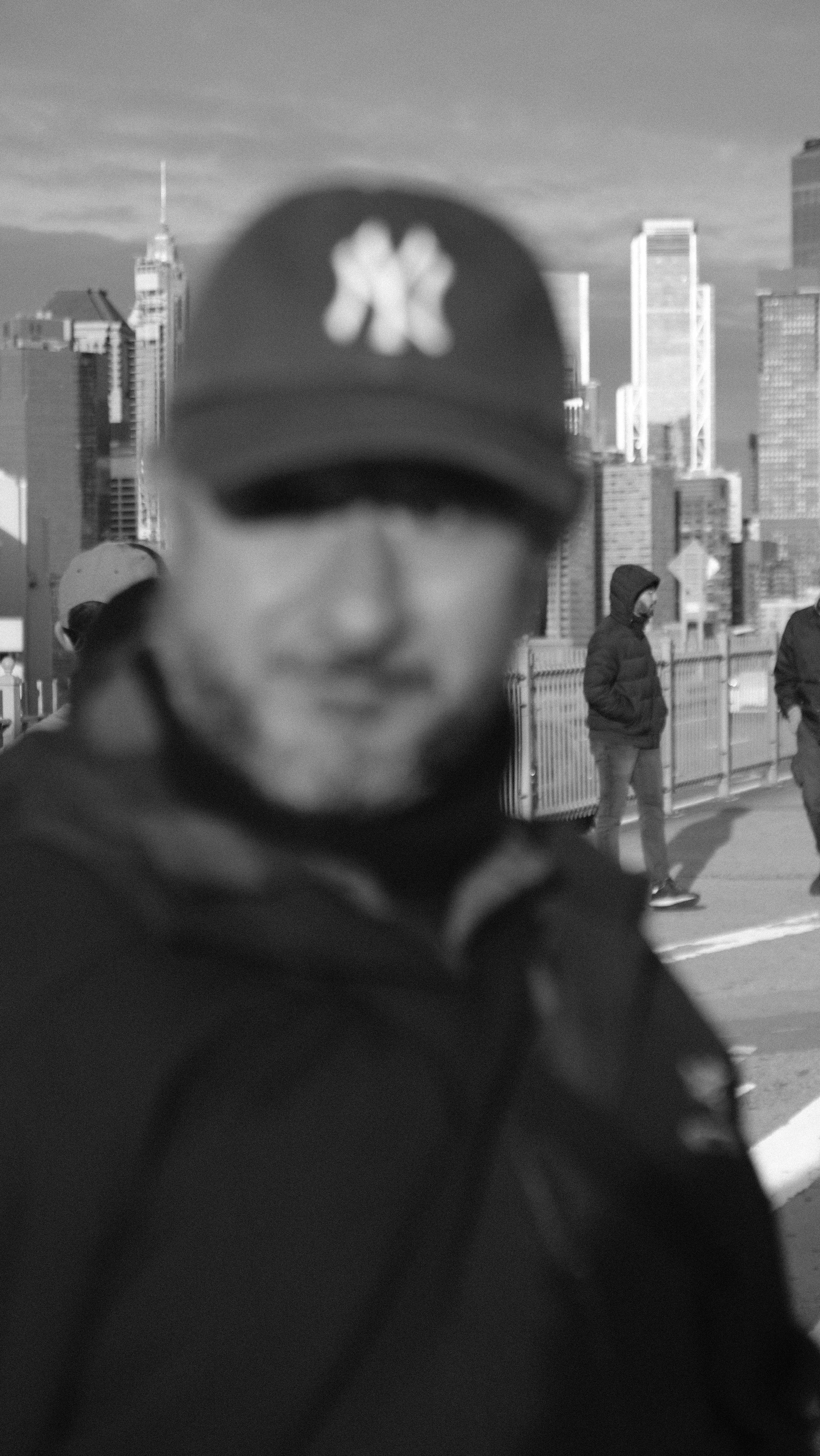 A person wearing a cap with a blurred logo, standing outdoors in front of a city skyline with skyscrapers, several other people in jackets, and a fence in the background.