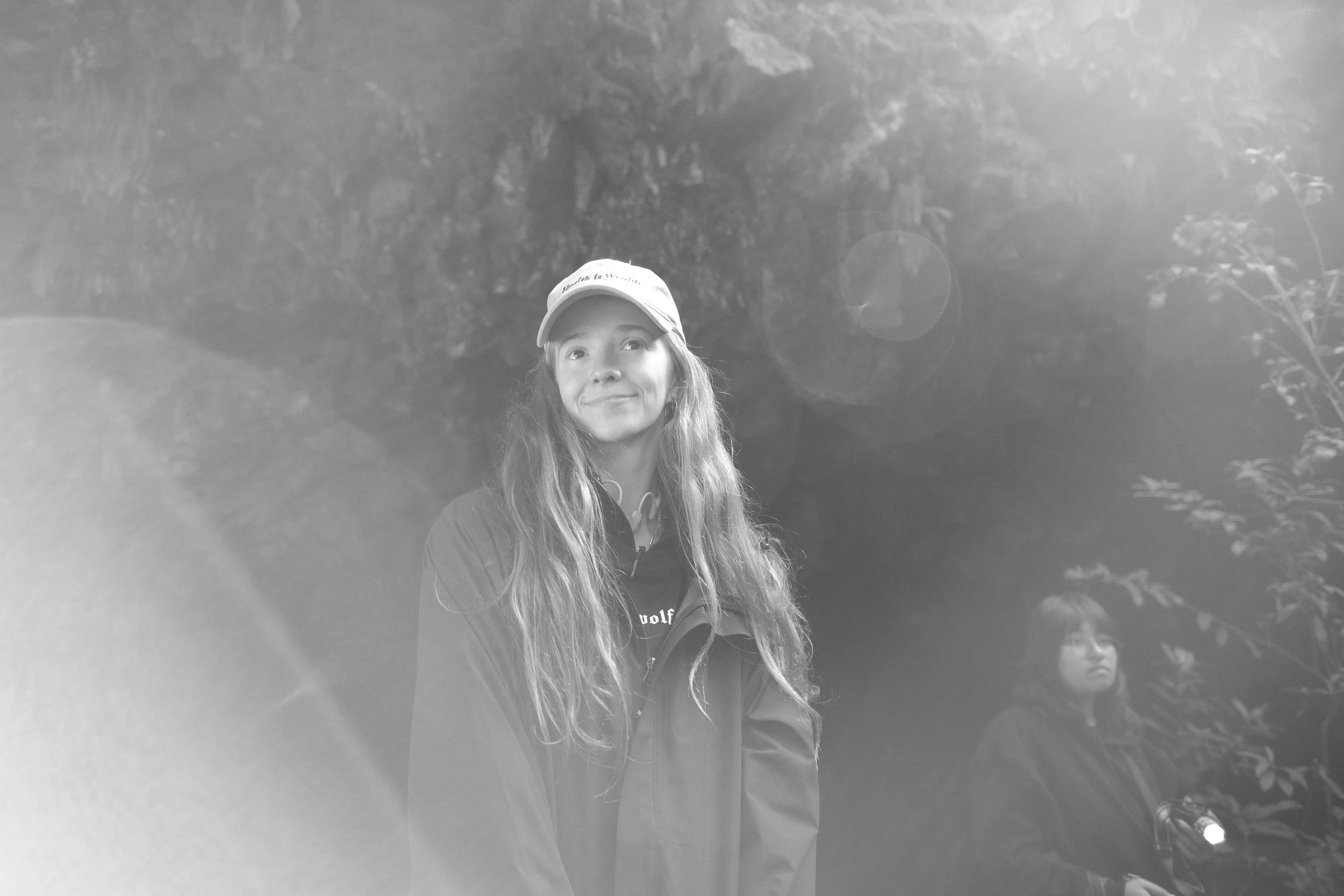Two young women standing outdoors at night, one wearing a cap and jacket, and the other holding a flashlight, with trees and foliage in the background.