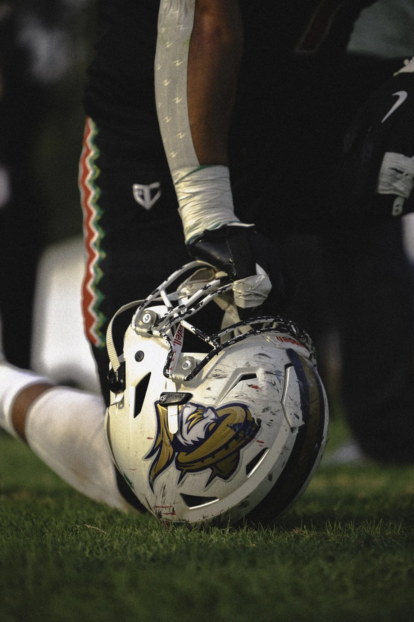 A football player kneeling on the field, holding a helmet with a lion logo, wearing a black uniform with an Italian flag stripe on the side of his pants and showing visible scratches and wear.