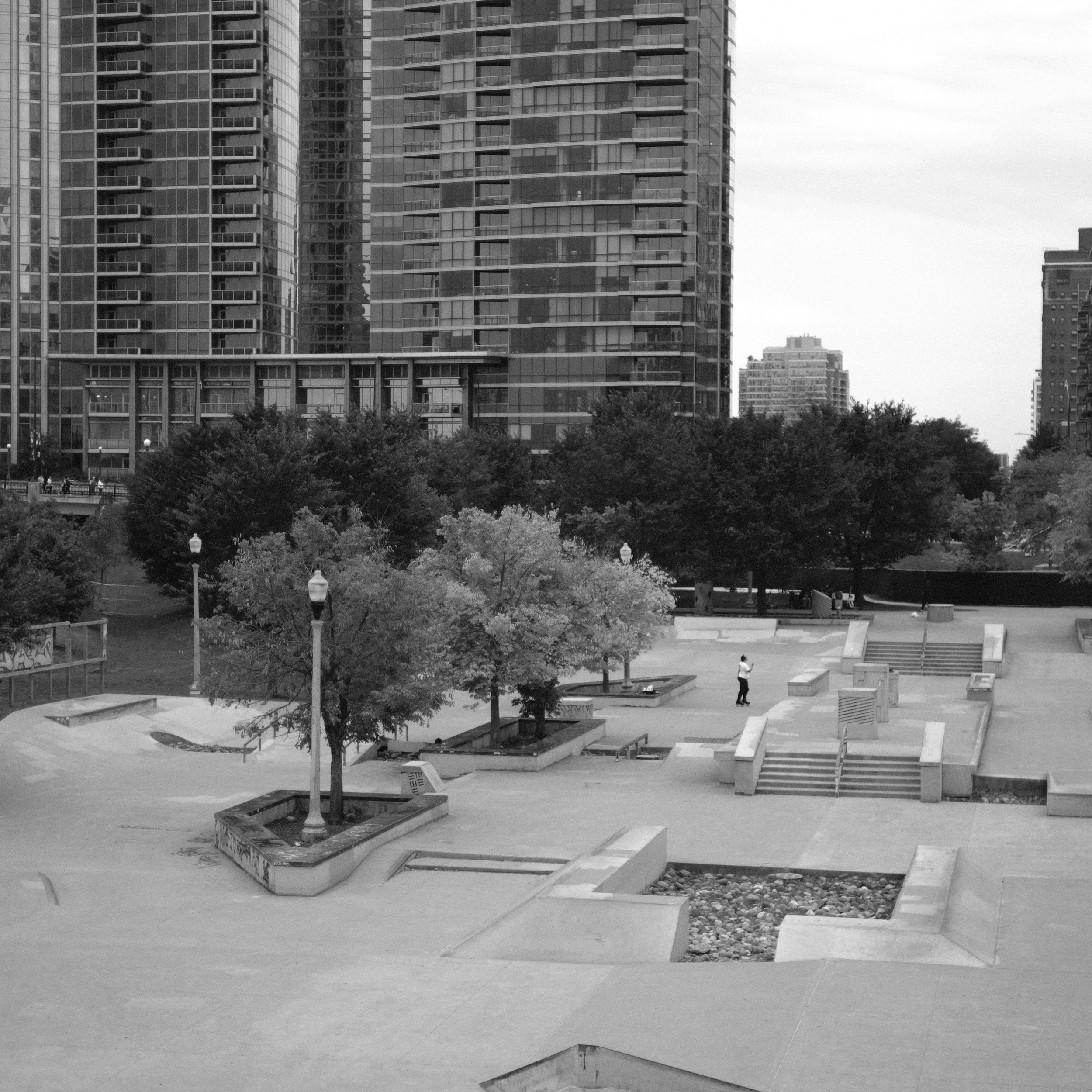 Black and white photo of an urban park with trees, benches, stairs, and people, surrounded by tall modern buildings.