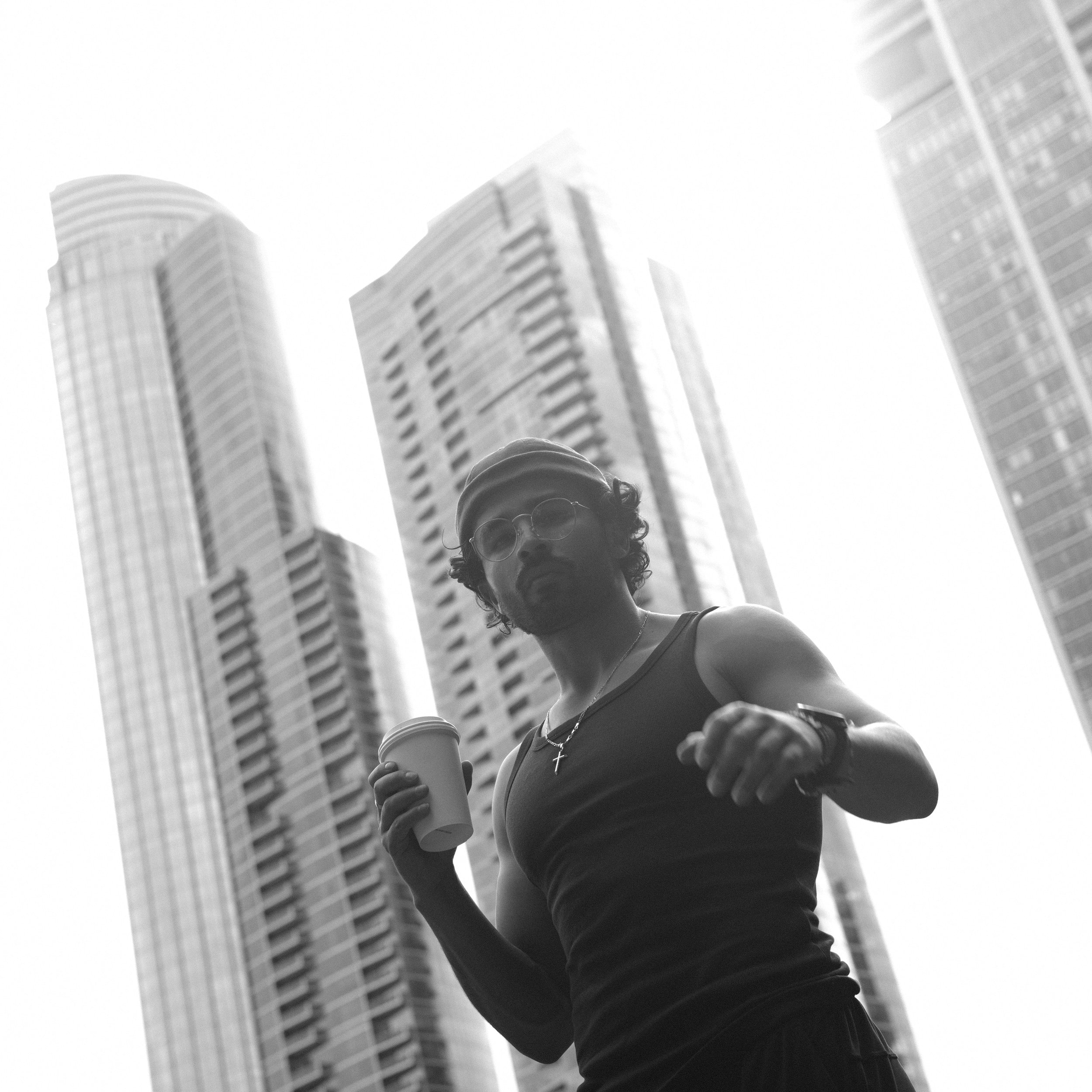 A man running in an urban area with tall skyscrapers in the background, holding a coffee cup, wearing sunglasses, a tank top, a watch, and a cross necklace, captured in black and white.