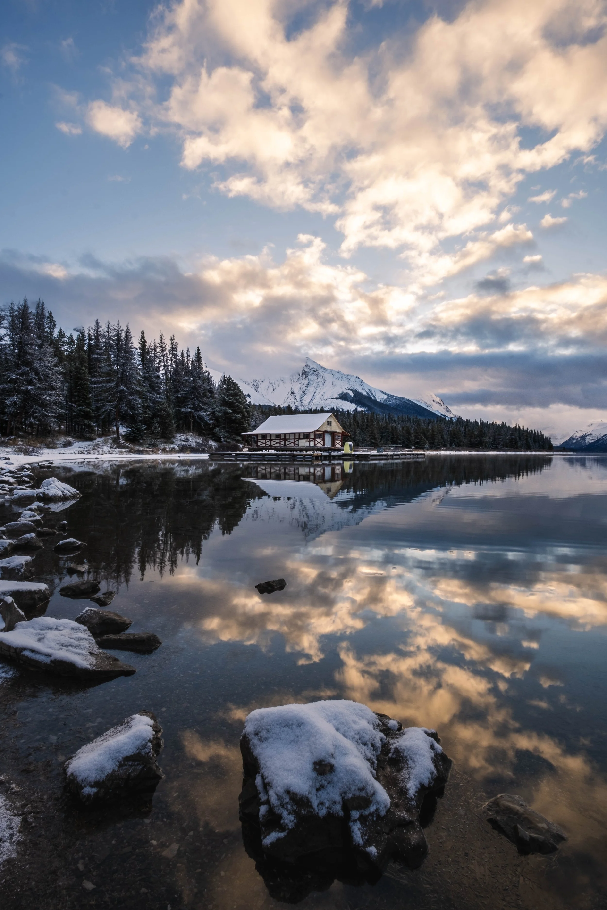 A snowy landscape featuring a calm lake reflecting a partly cloudy sky, snow-covered rocks in the foreground, a forested shoreline, a house on a pier, and snow-capped mountains in the background.