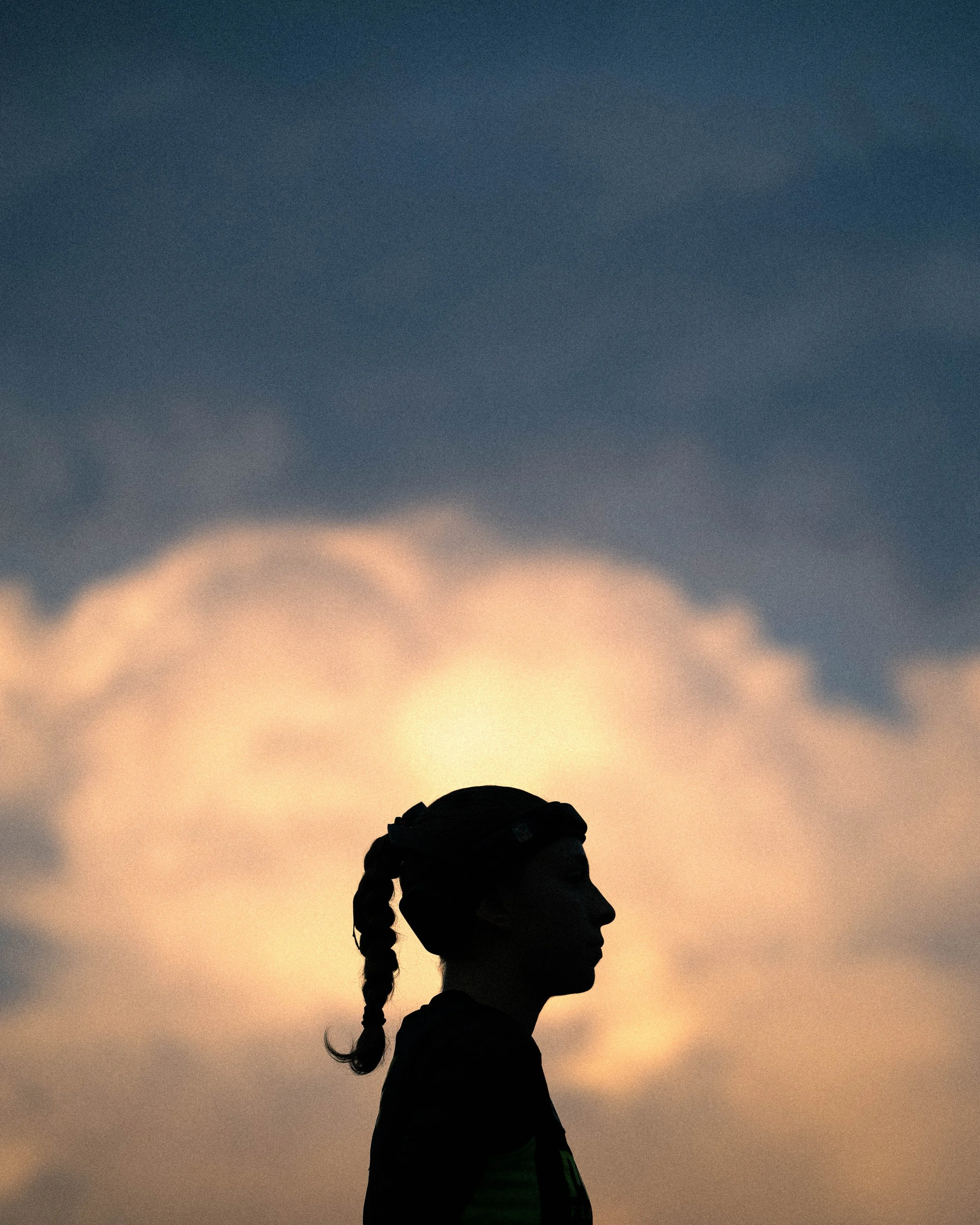 Silhouette of a girl with braided hair and a cap against a cloudy sky at sunset.