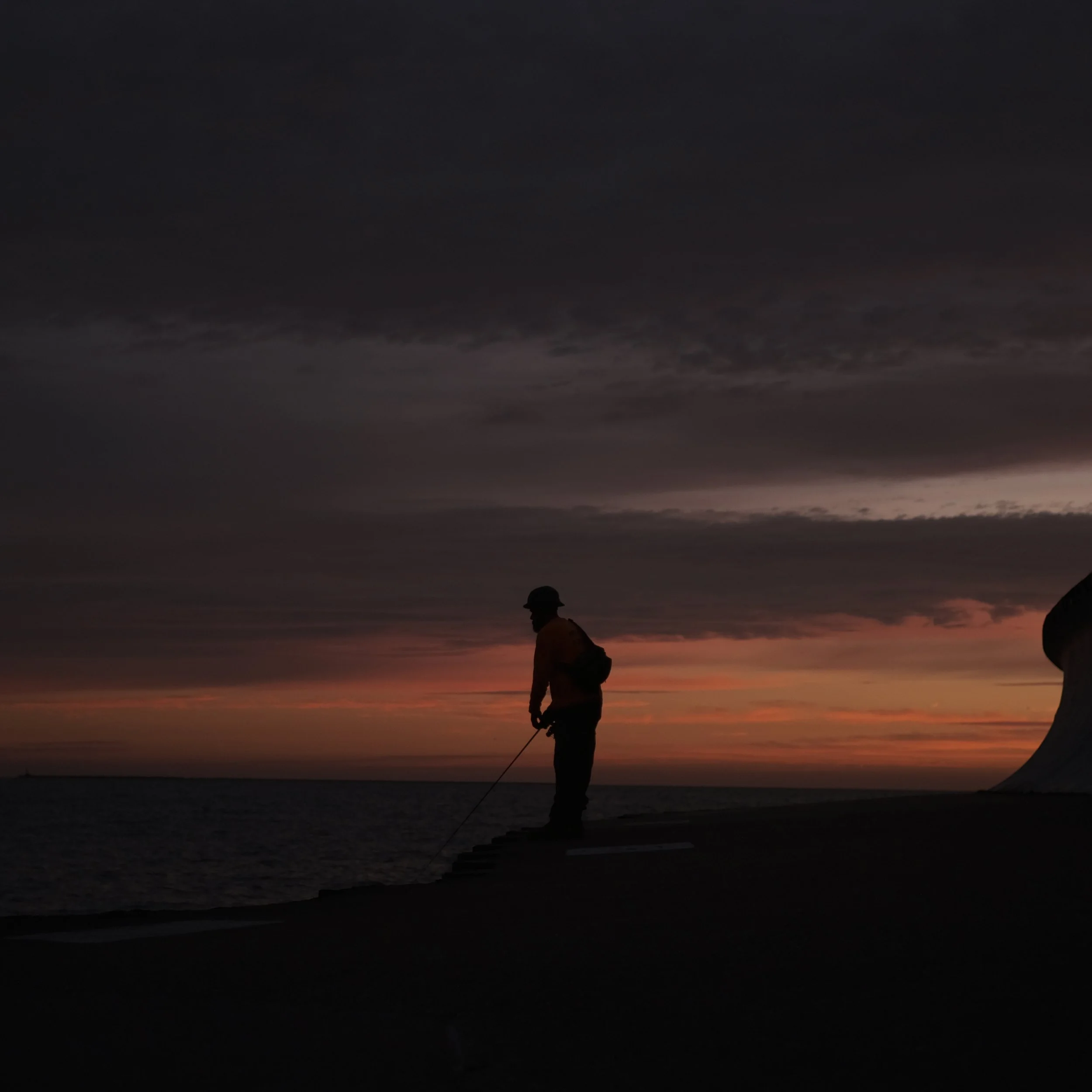Silhouette of a person fishing by the water at sunset with a dramatic cloudy sky in the background.