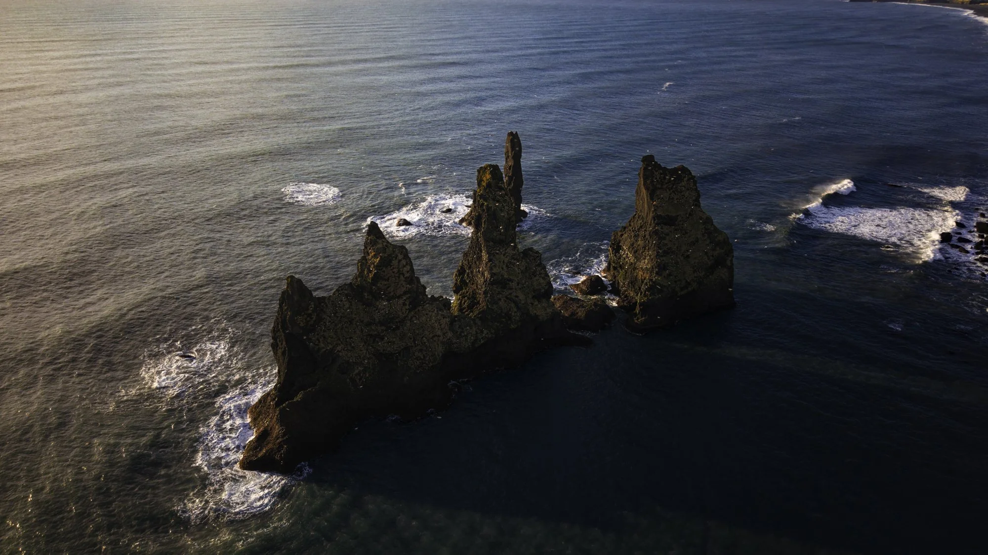 Aerial view of three large dark rocky sea stacks rising from the ocean, with waves crashing at their bases, under a clear sky.