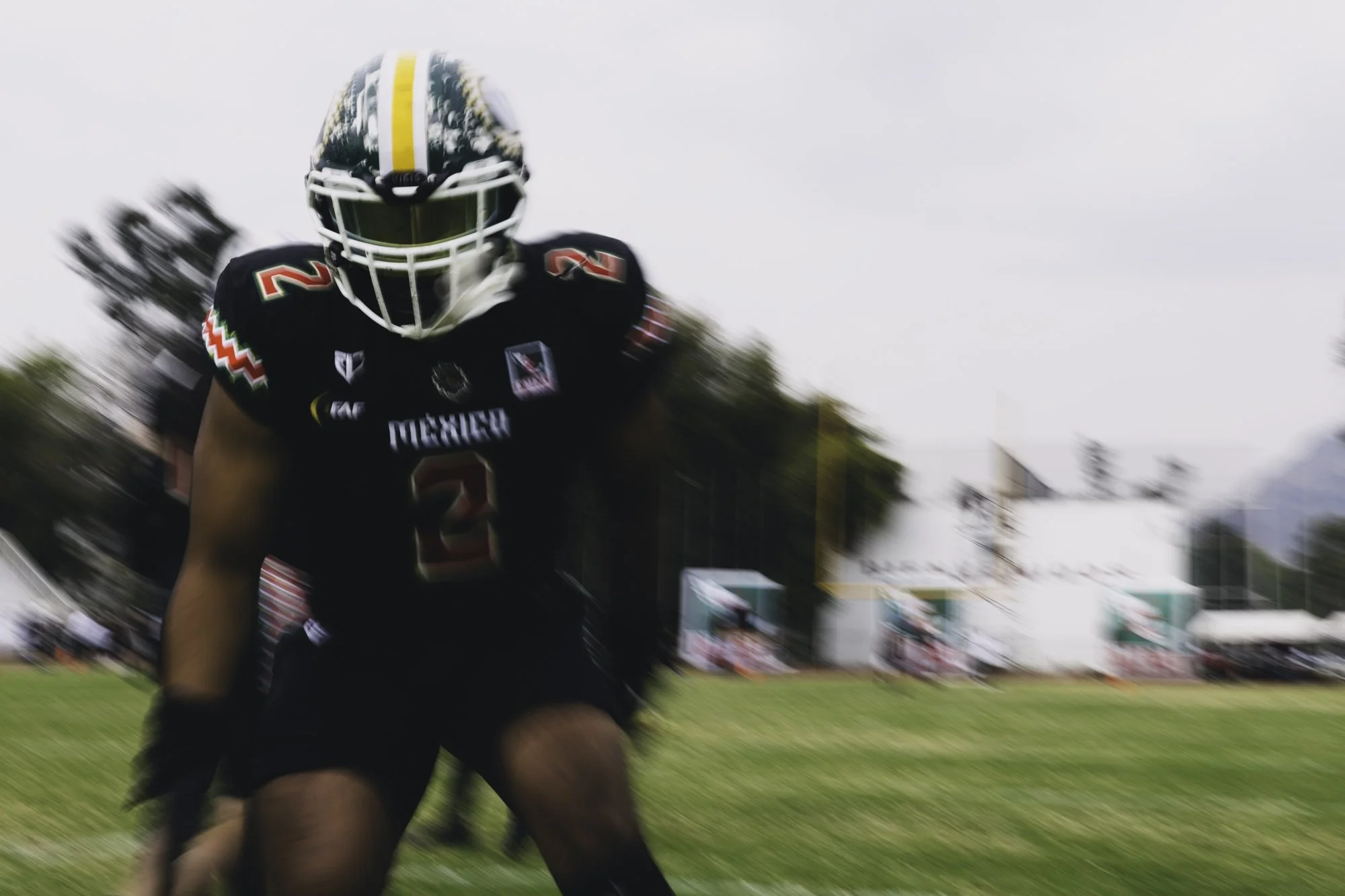 A football player in a black uniform with the number 2 running on a field, wearing a helmet with a yellow and green stripe, and the background is slightly blurred.