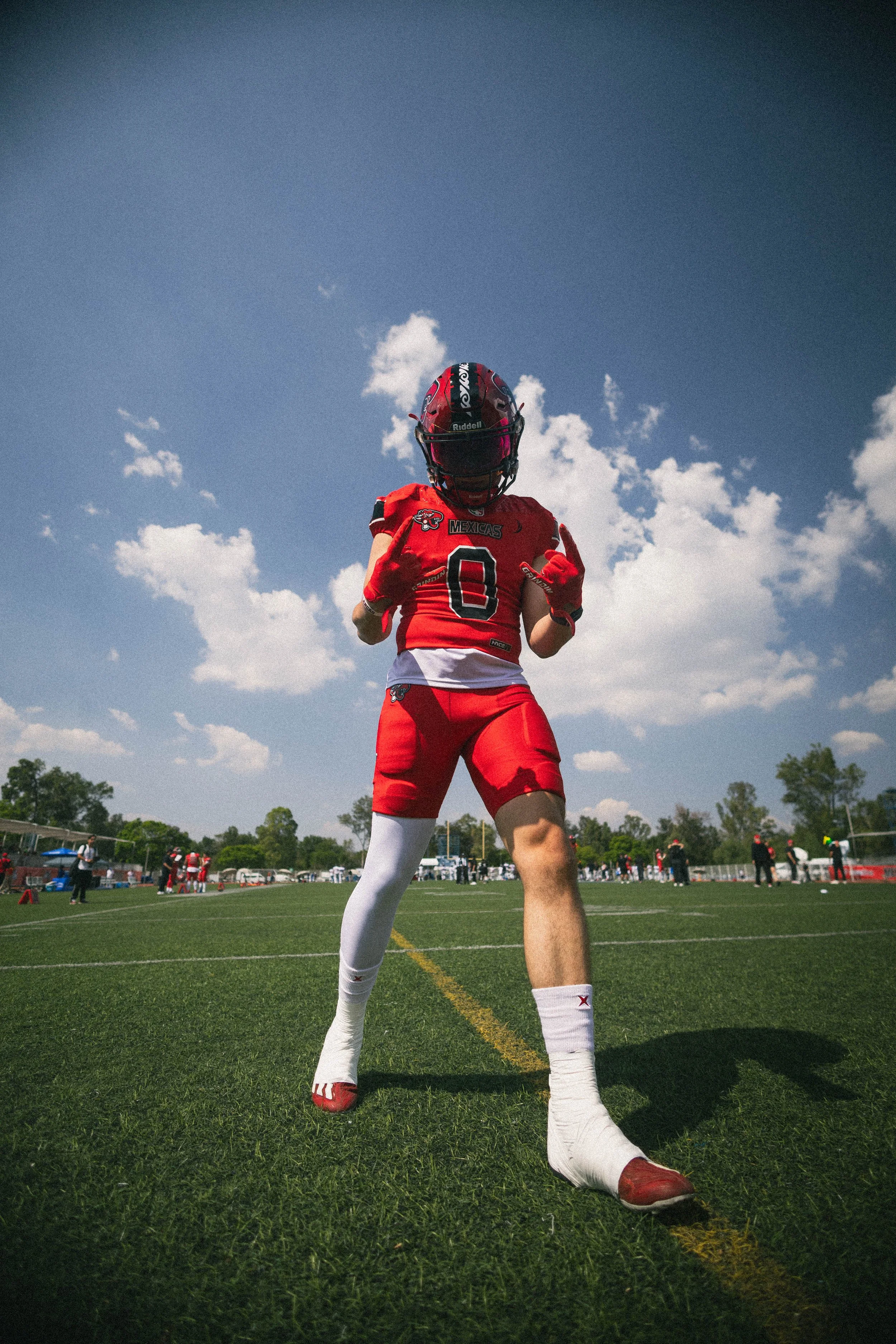 A football player stands on the field wearing a red uniform with the number 0, a red helmet, white socks, and red shoes, making a gesture with both hands against a bright blue sky.