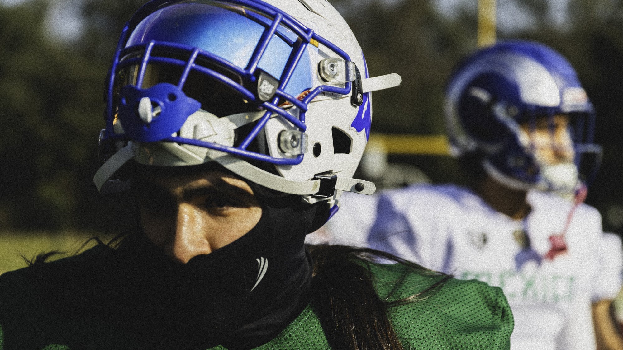 A football player wearing a white and green uniform with a black face mask and a helmet with a blue faceguard, standing outdoors.