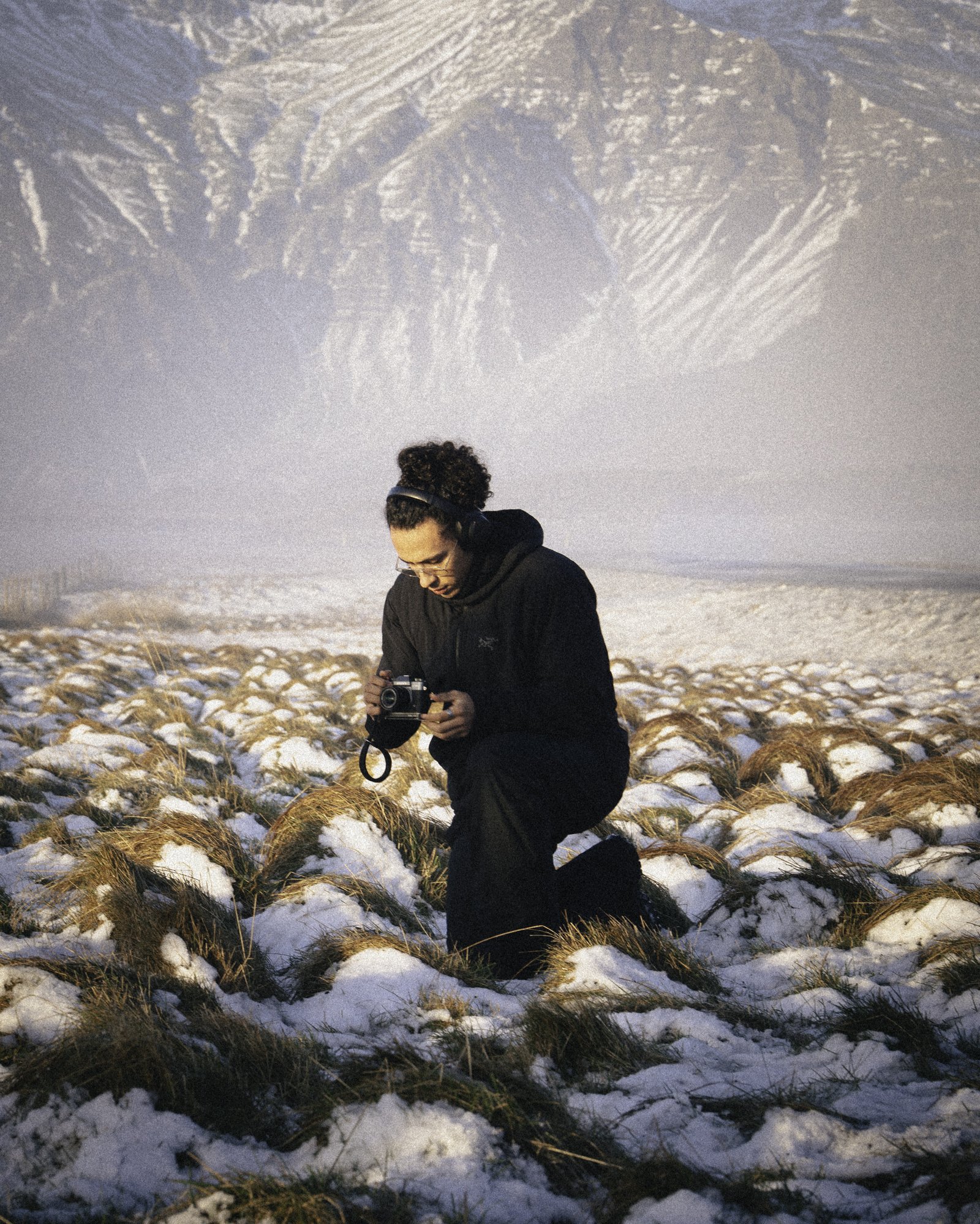A person with curly hair wearing headphones and a black jacket is kneeling on snow-covered ground while looking at a camera in their hands, with snow-covered grass around them and a foggy mountain in the background.