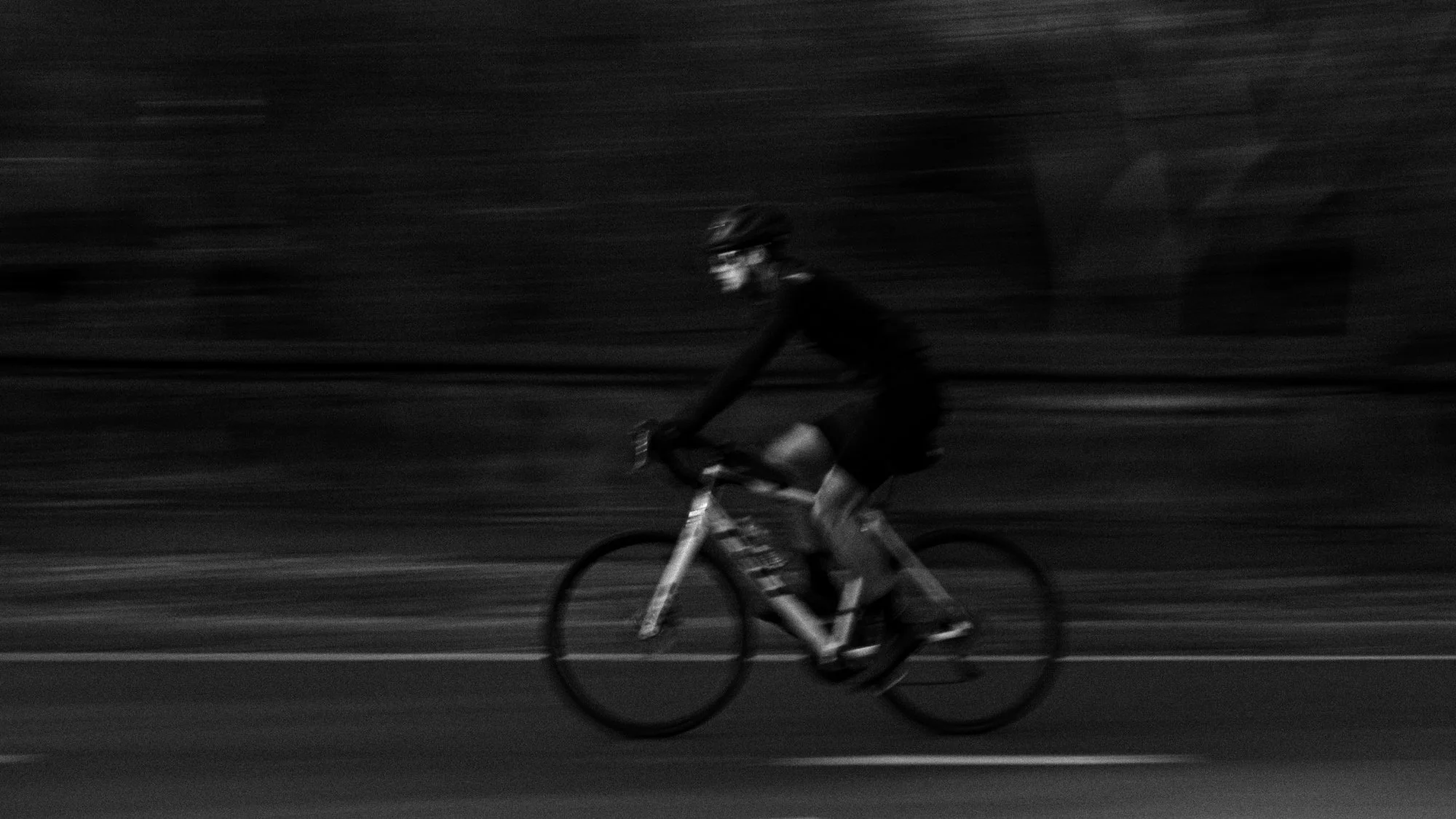 A person riding a bicycle on a road at night, wearing a helmet and dark clothing, with a blurred background indicating motion.