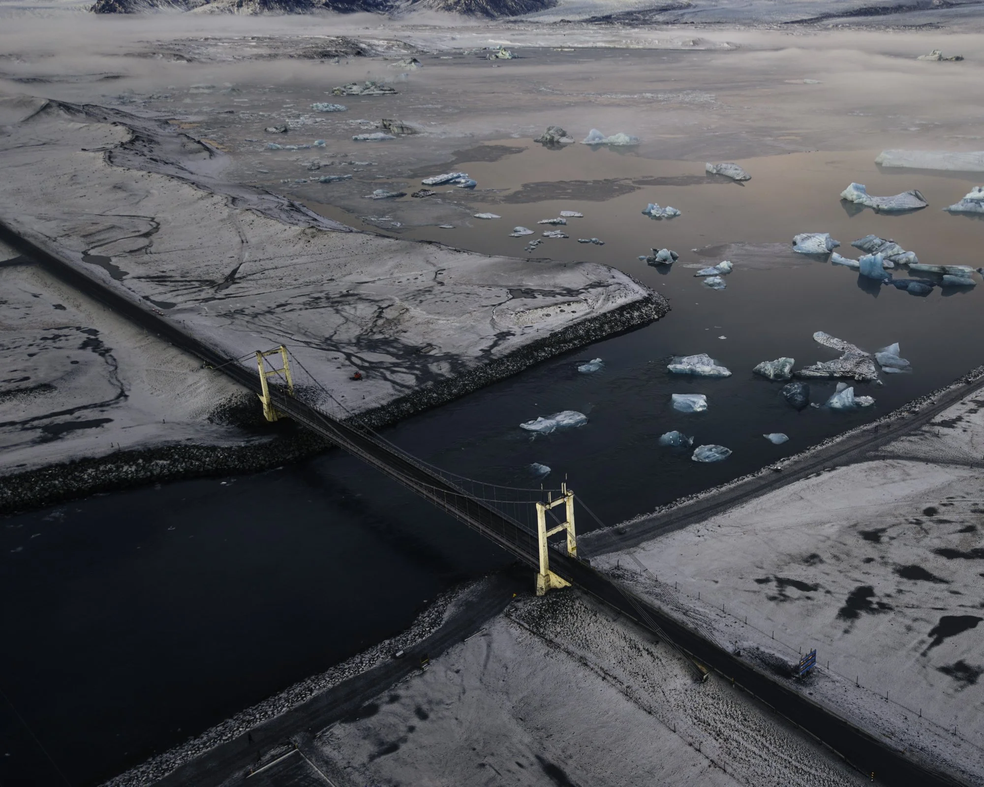 Aerial view of a snow-covered bridge crossing a waterway with floating icebergs and snow-covered land around.