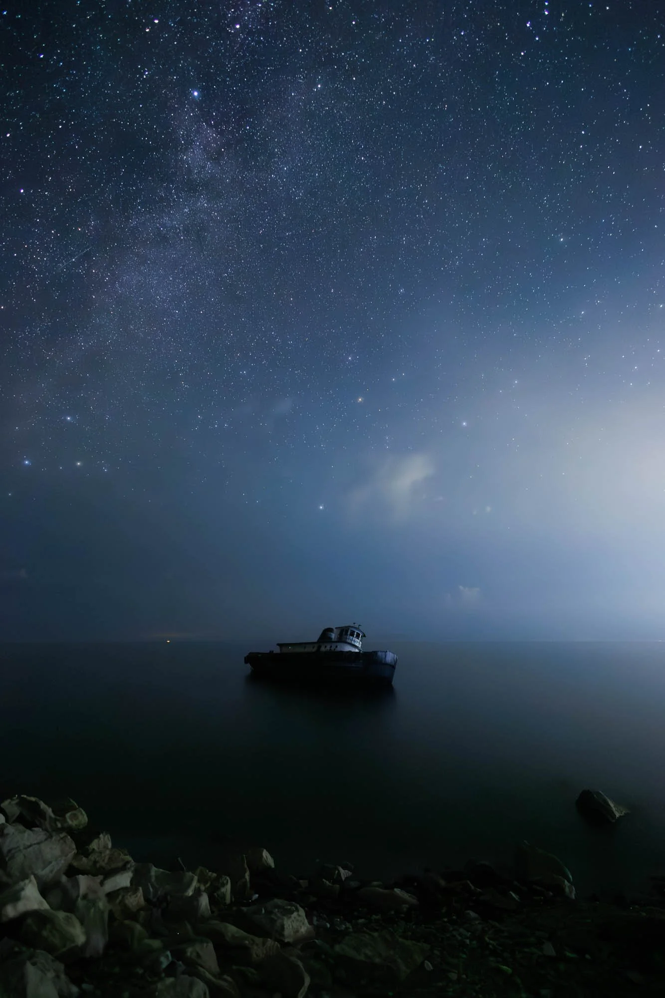 A nighttime scene of a boat floating on calm water under a star-filled sky with the Milky Way visible.