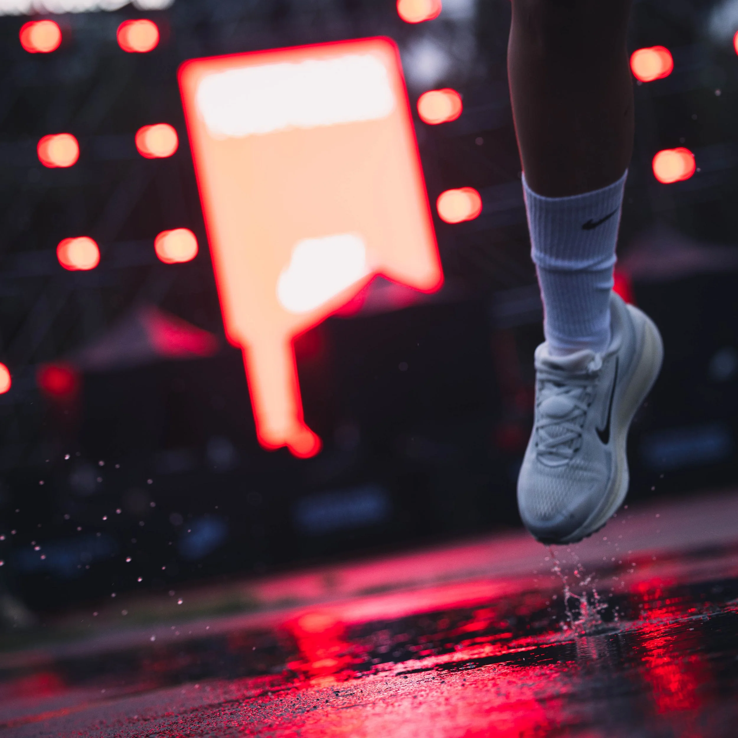 Close-up of a runner's leg and white Nike sneaker with a black swoosh, running on wet pavement during evening with illuminated red digital display in background.