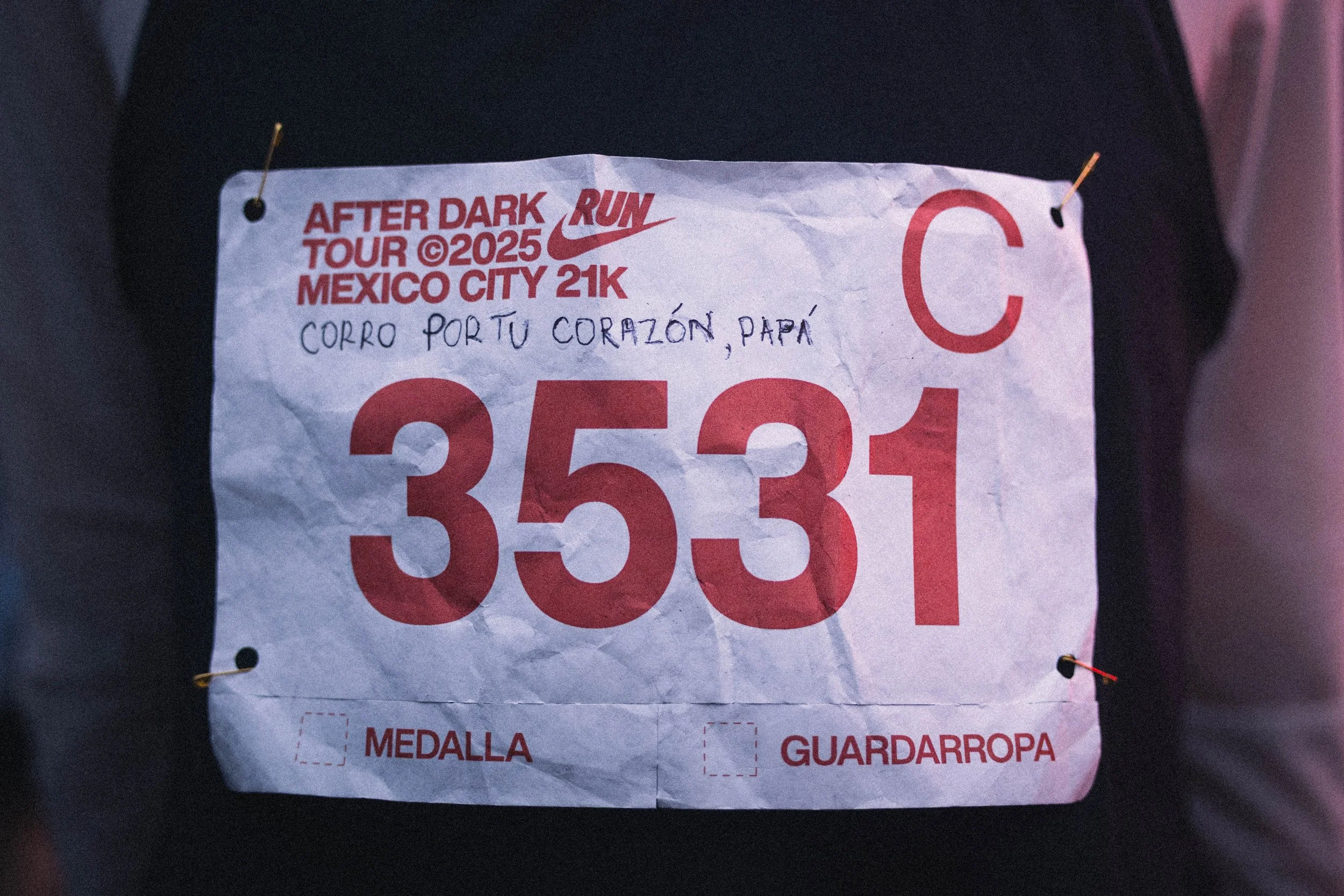 Race bib pinned to a person's black shirt with the text 'After Dark Run Tour 2025 Mexico City 21K'. The bib has large red numerals '3531'. It includes handwriting that says 'CORRO POR TU CORAZÓN PAPÁ'. There are two small checkboxes labeled 'Medalla'