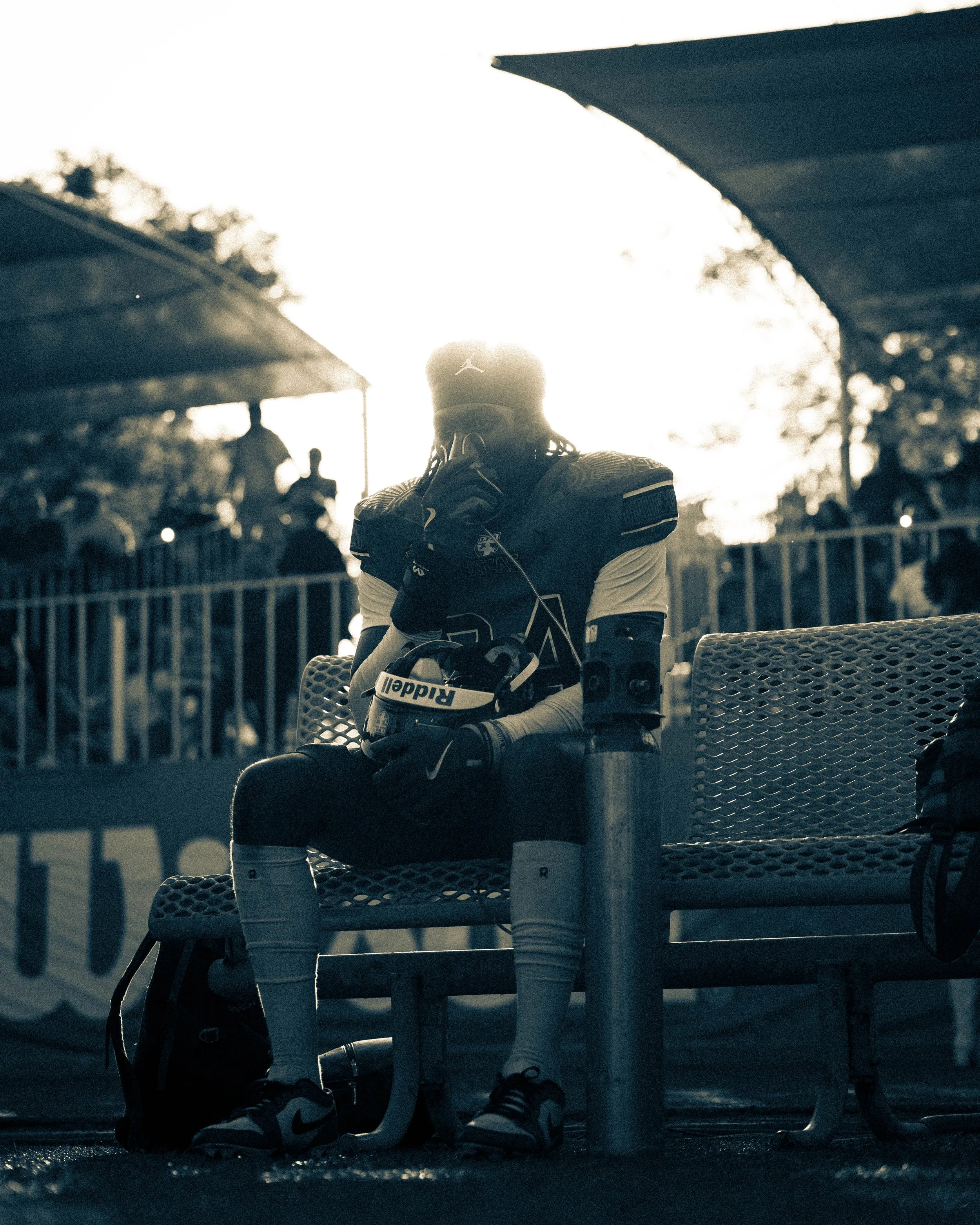 A football player sitting on a bench during sunset, wearing football gear, holding a helmet, and drinking from a water bottle.
