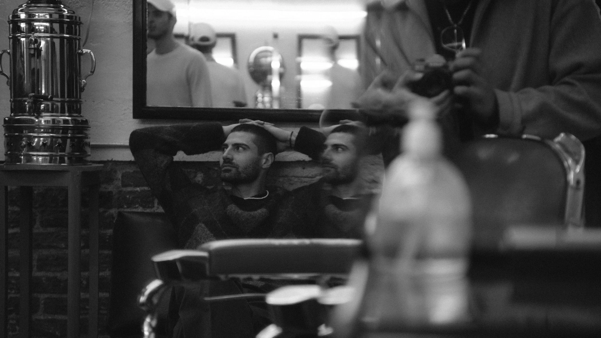 A man sitting on a couch with his hands behind his head, gazing contemplatively to the side. There is a mirror behind him reflecting other people in a room. A person on the right is holding a dog. The scene is in a cafe or restaurant setting, with va