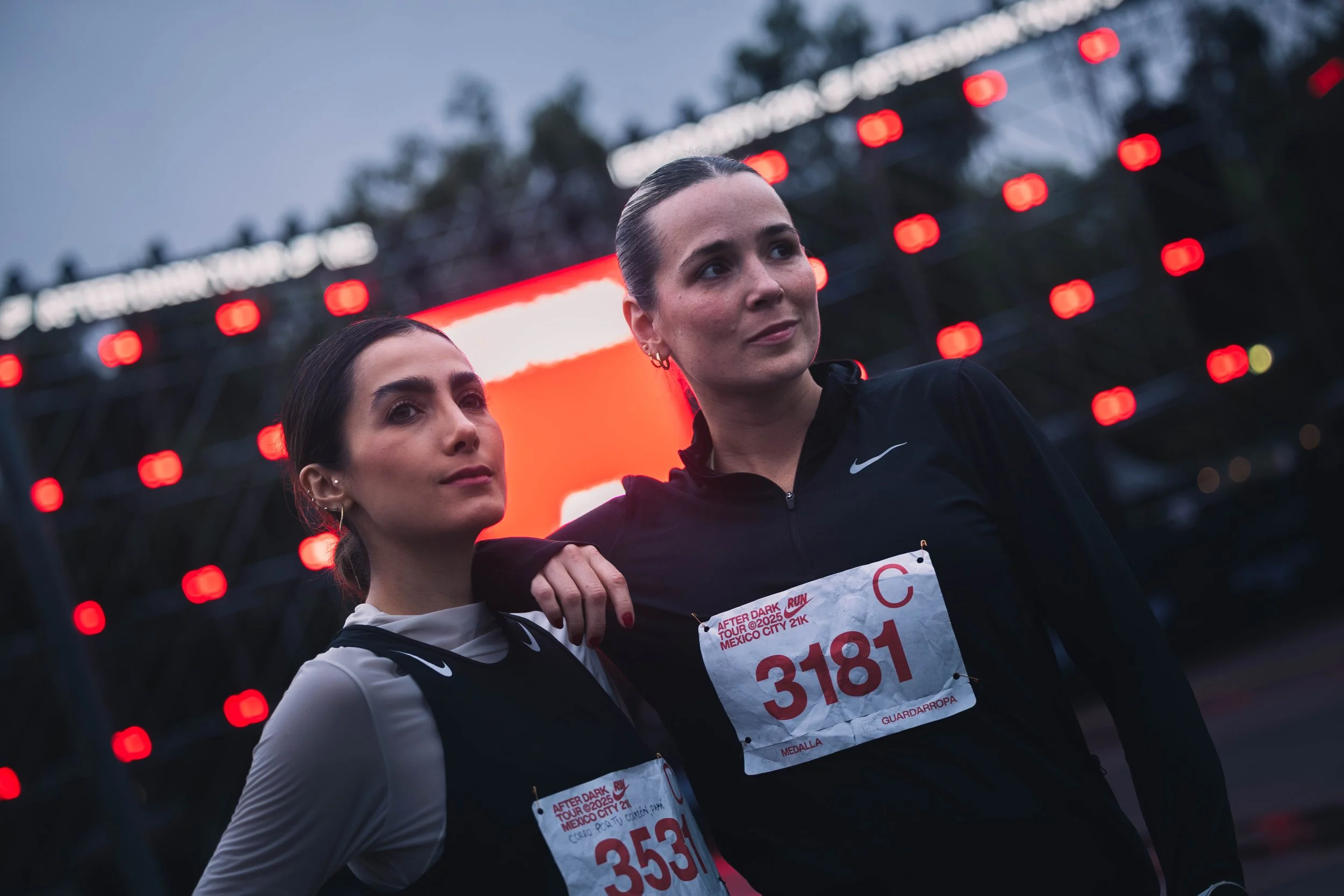 Two women with race bibs, posing together during a running event at dusk with a large illuminated screen behind them.