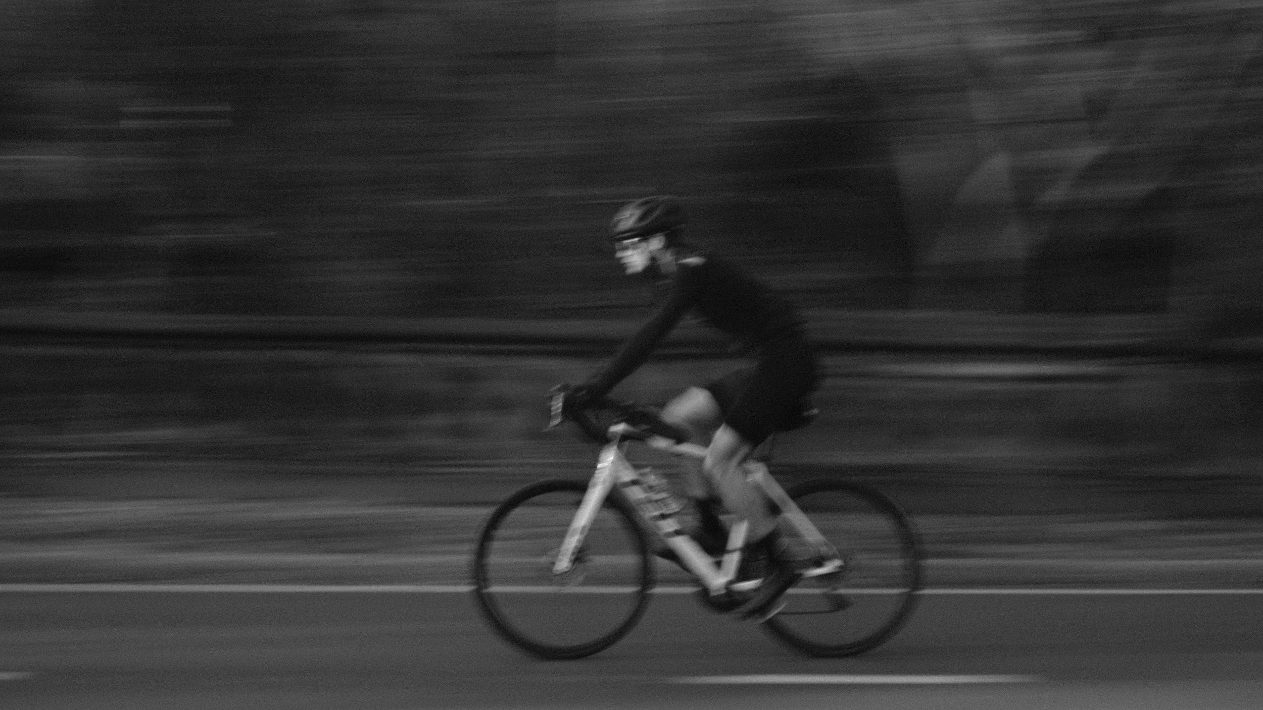 A person riding a bicycle on a road, wearing a helmet and dark clothing, captured in motion with a blurred background.