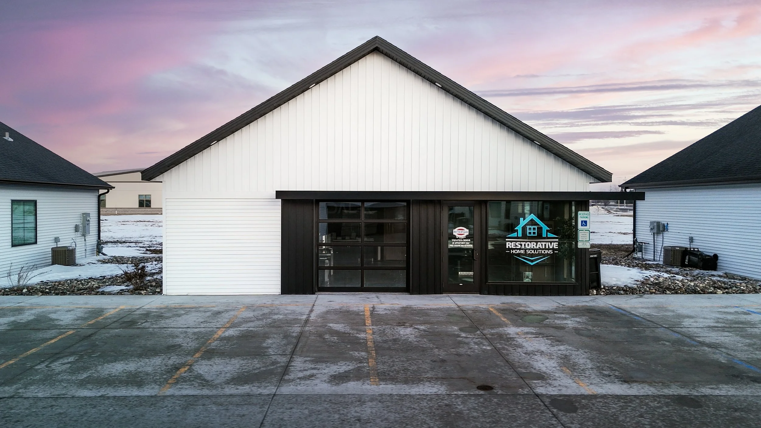 Front of a small business building with signs for 'Restorative Home Solutions' and parking spaces, with snow on the ground and a colorful sunset sky.