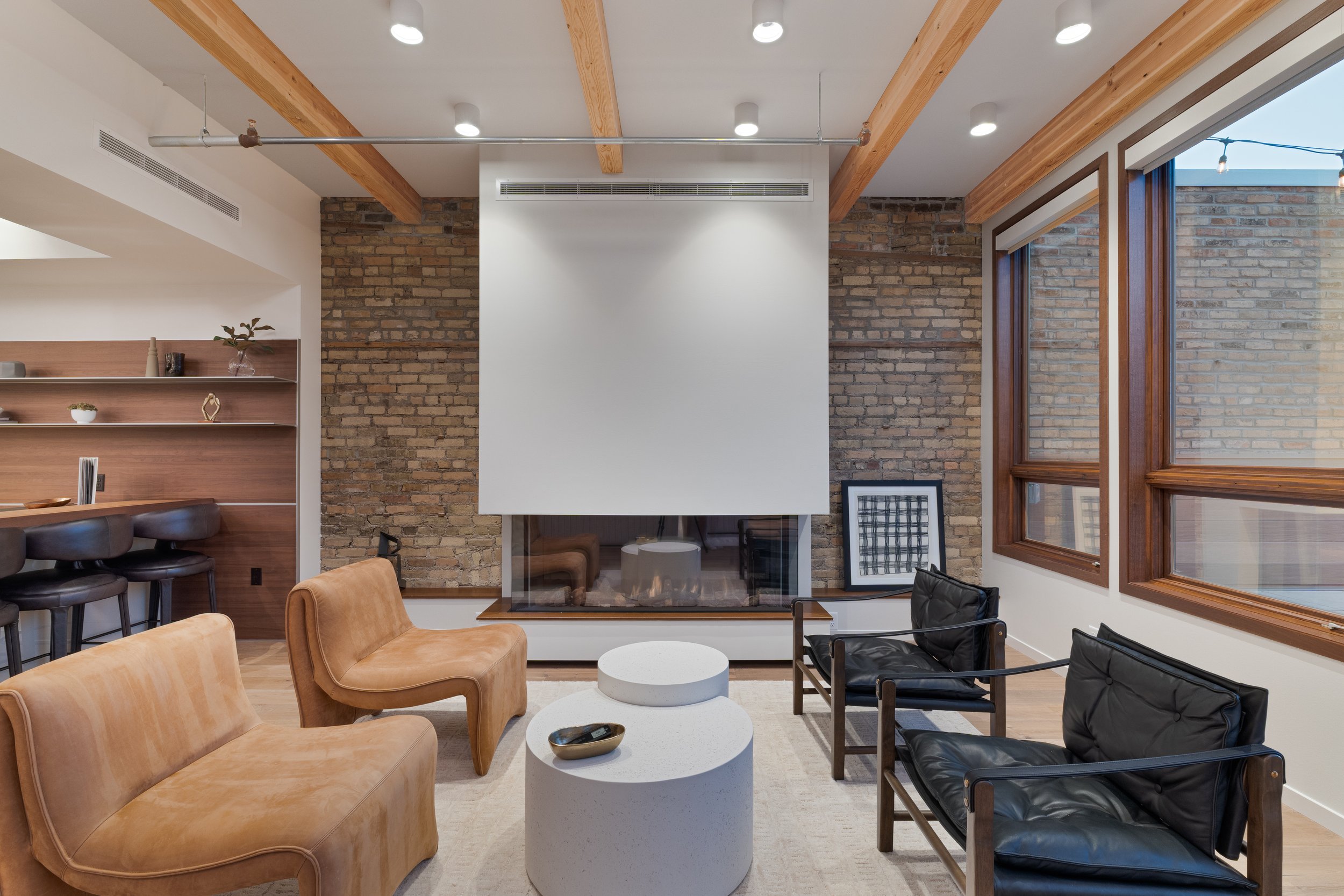 Modern living room with a brick fireplace, beige armchair, black chairs, large windows, and wooden accents.