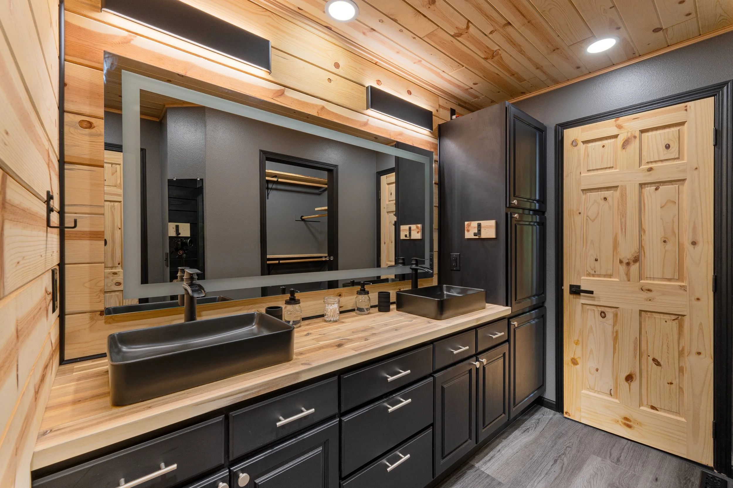 Bathroom with a large mirror, black sinks, black cabinets, light wood countertop, wooden walls, and a wooden door.
