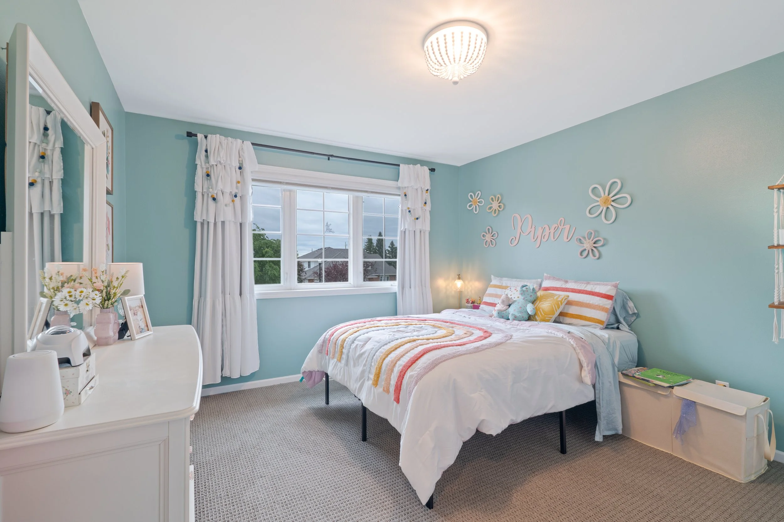 A bedroom with pastel blue walls, a white bed with striped pillows, and a wall decoration that says 'Paper' with flower designs. The room has a window with white curtains, a white dresser with flowers and picture frames, and a beige carpet.