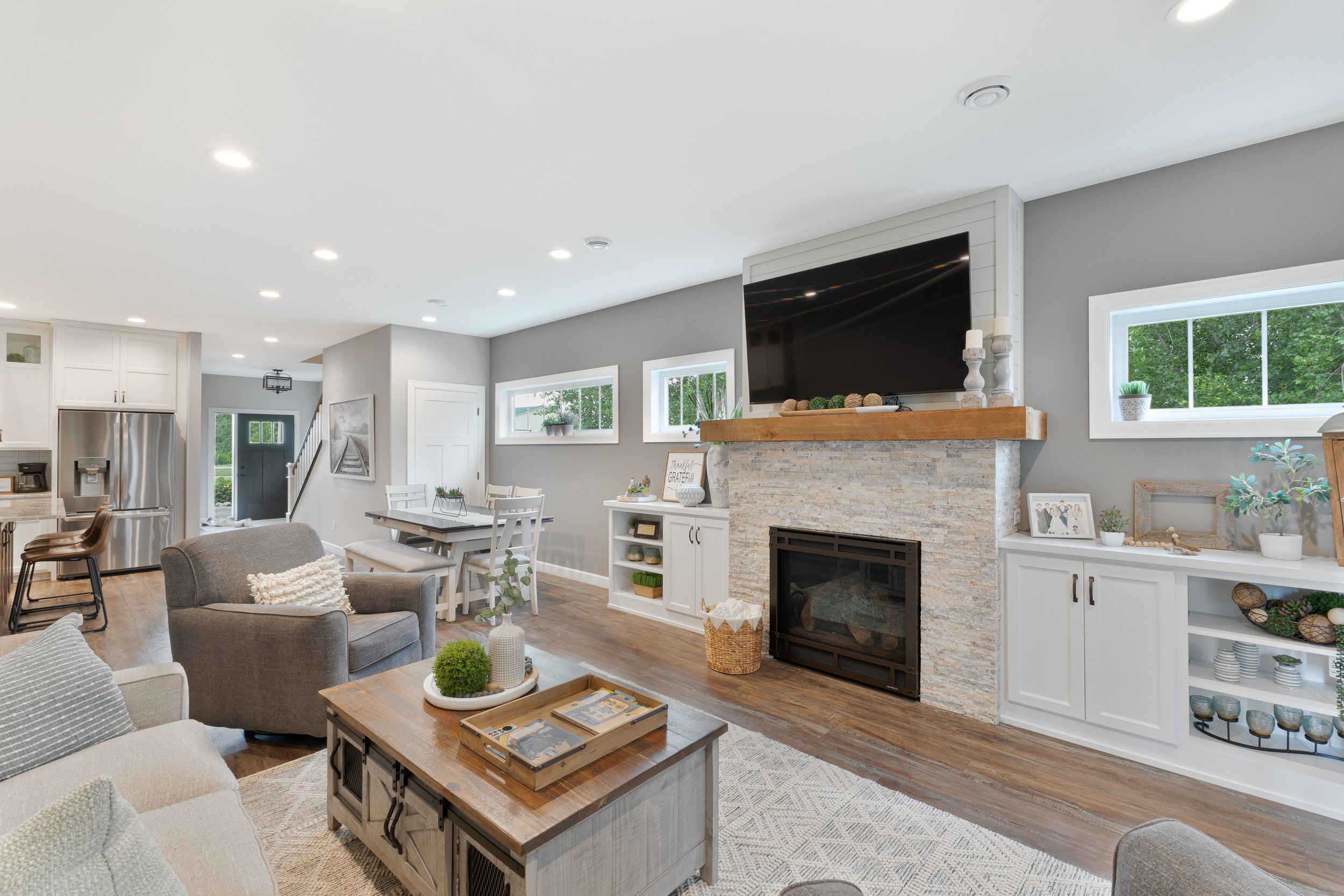 Living room with gray walls, white built-in cabinets, a fireplace, a large flat-screen TV mounted above it, and a mix of gray, white, and beige furniture. Multiple windows provide natural light, and decorative items are arranged on surfaces.