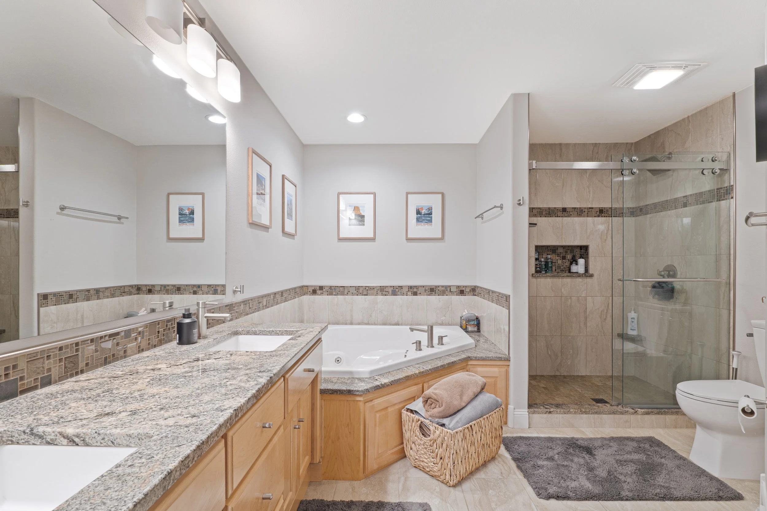 Modern bathroom featuring a double sink granite countertop, corner bathtub, glass-enclosed shower, framed art on the wall, and a basket of towels on the floor.