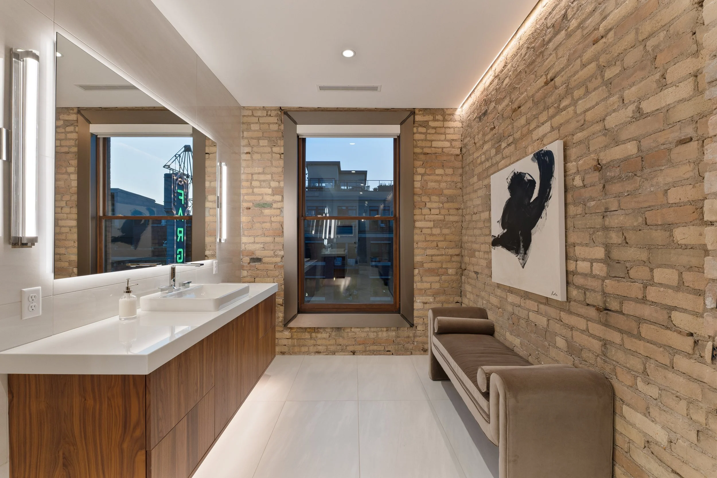 Modern bathroom with exposed brick walls, a large mirror, a white sink and wooden vanity, window showing building and street view, and an armchair against the brick wall with art hanging above.