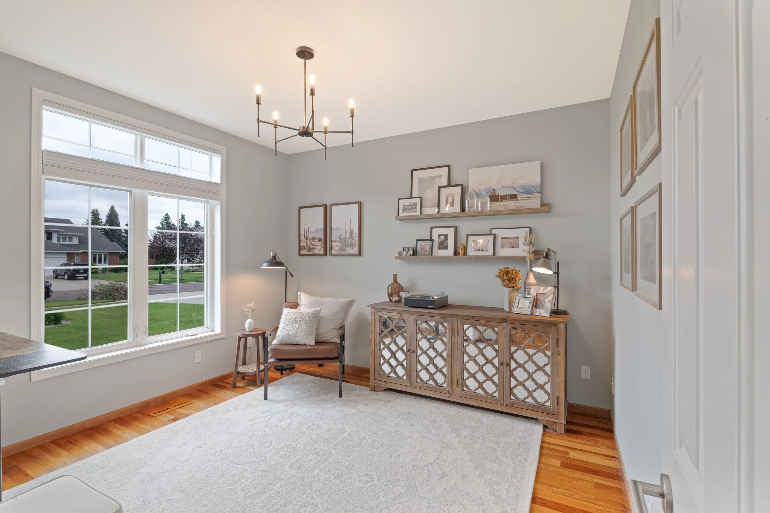 Living room with a large window, gray walls, a wooden sideboard, wall art, and a single armchair with a side lamp.