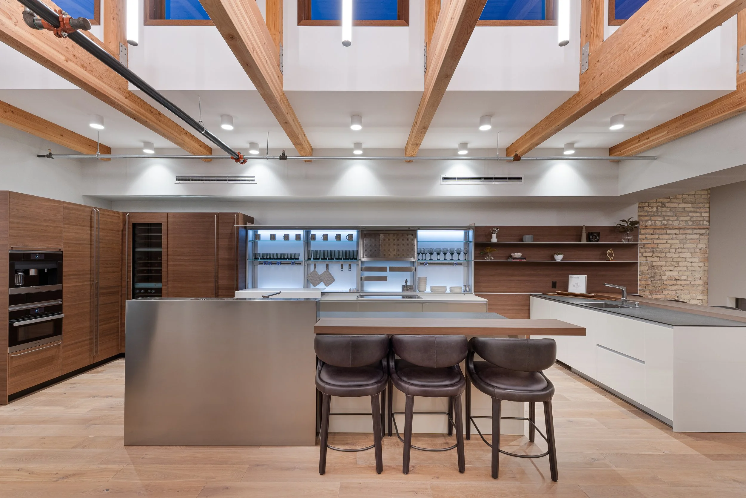 Modern kitchen with wooden beams on the ceiling, white walls, and a mix of wood and white cabinetry. The kitchen features a central island with a gray countertop, dark brown bar stools, and open shelves with dishes and decor. There is a brick wall se